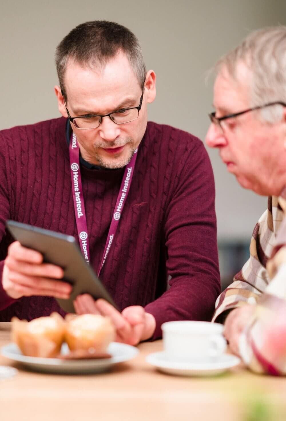 Two men seated at a table, one showing the other a tablet. Cupcakes and a coffee cup are on the table. - Home Instead Poole