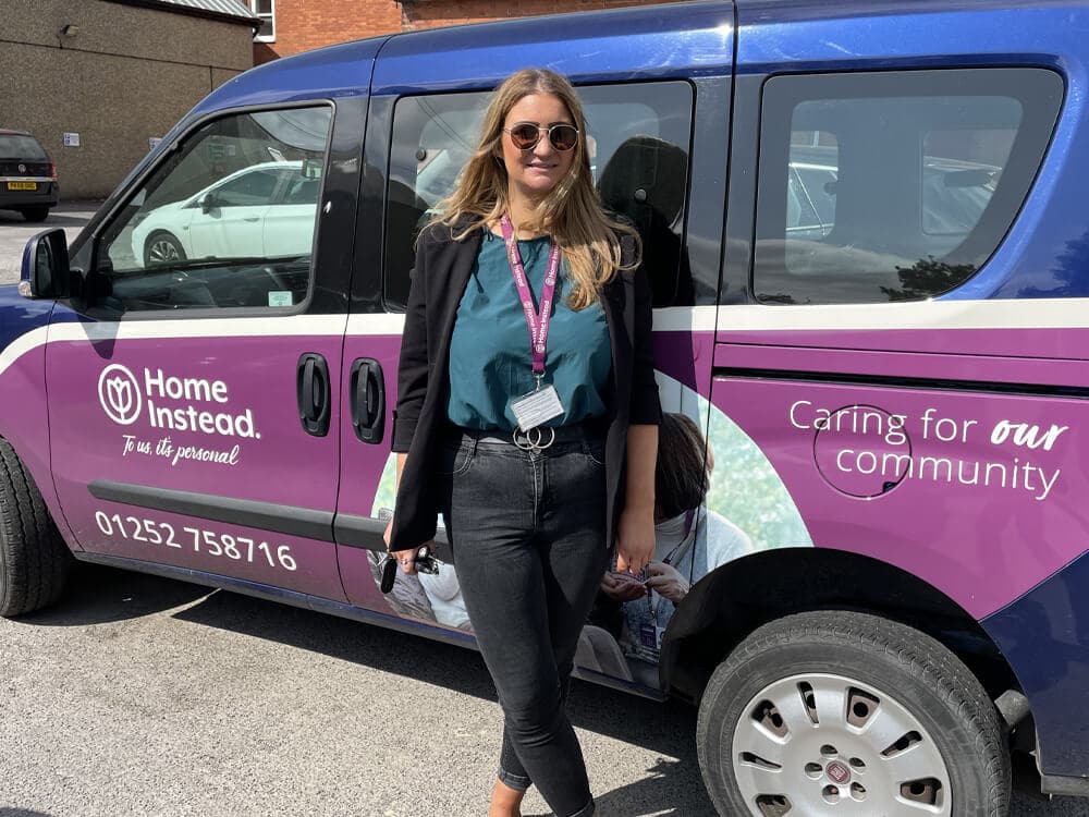 A woman in sunglasses stands in front of a Home Instead car with a "Caring for our community" slogan. - Home Instead