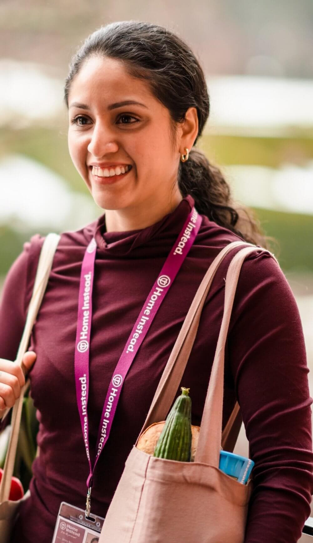 A woman smiles while carrying a tote bag filled with groceries, wearing a purple lanyard and a maroon top. - Home Instead