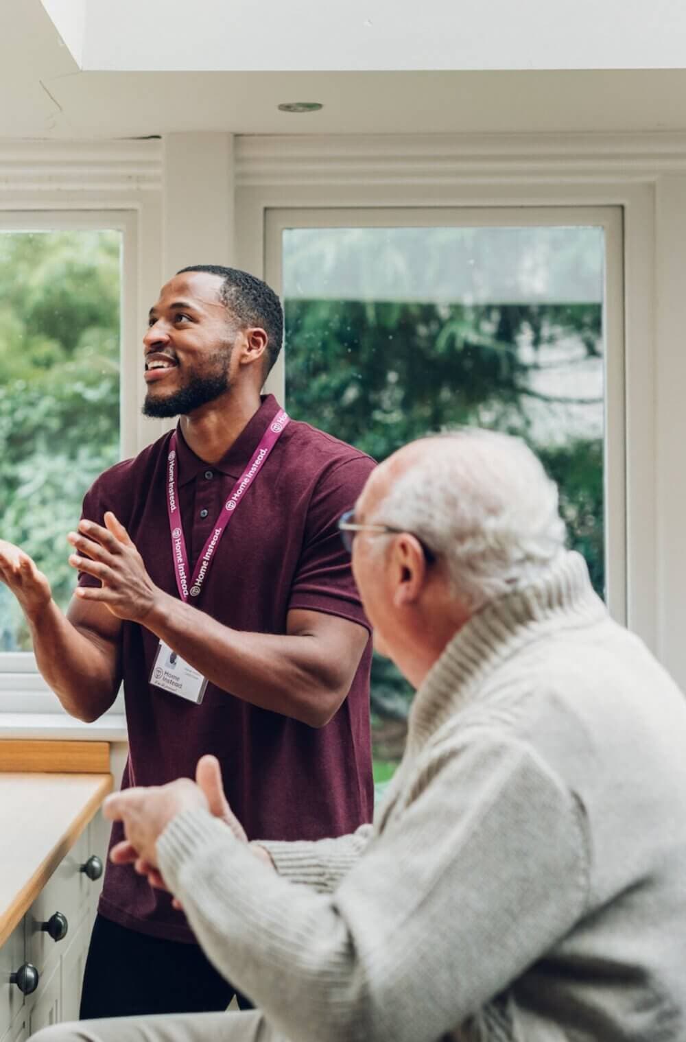 A young man in a maroon shirt converses with an elderly man in a grey sweater in a bright room with large windows. - Home Instead