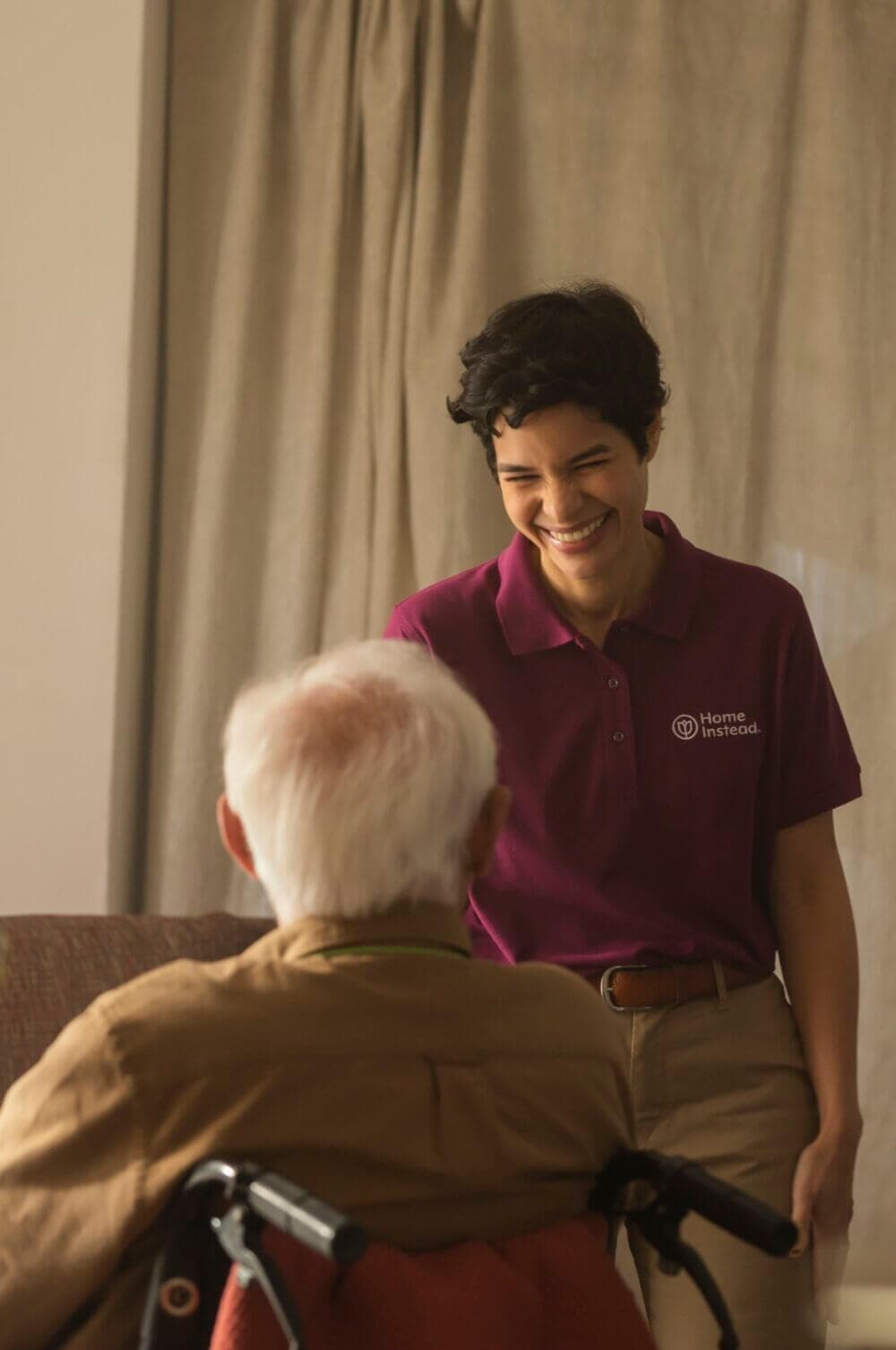 A caregiver smiles warmly at an elderly person sitting in a wheelchair. - Home Instead
