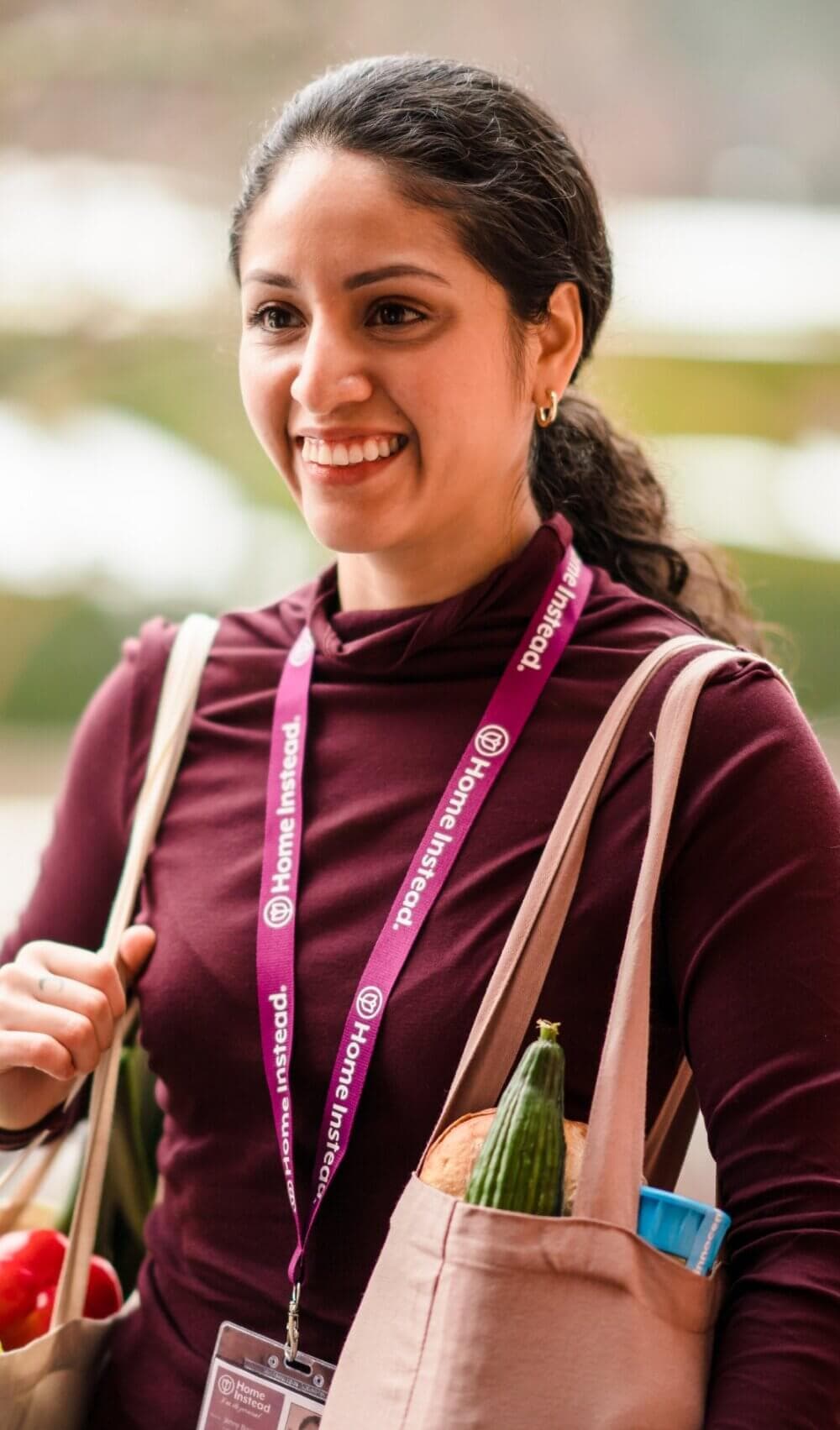 A smiling person carrying grocery bags and wearing a lanyard with an ID badge. - Home Instead Poole