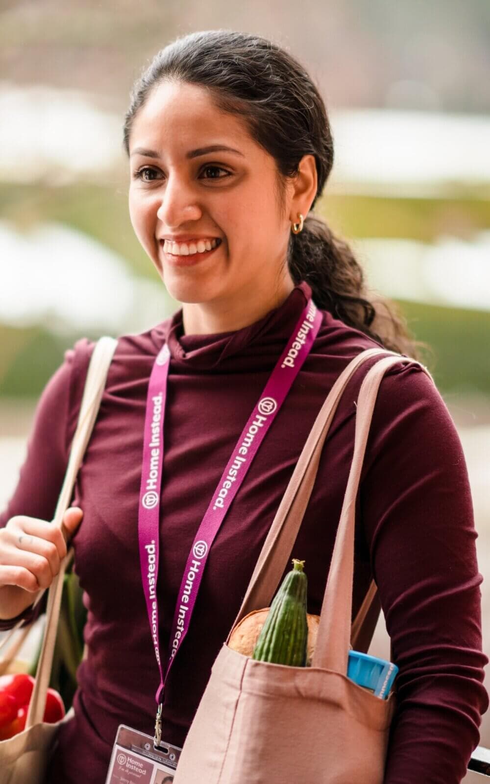 Smiling woman carrying grocery bags, wearing a maroon shirt and a lanyard around her neck. - Home Instead Southampton