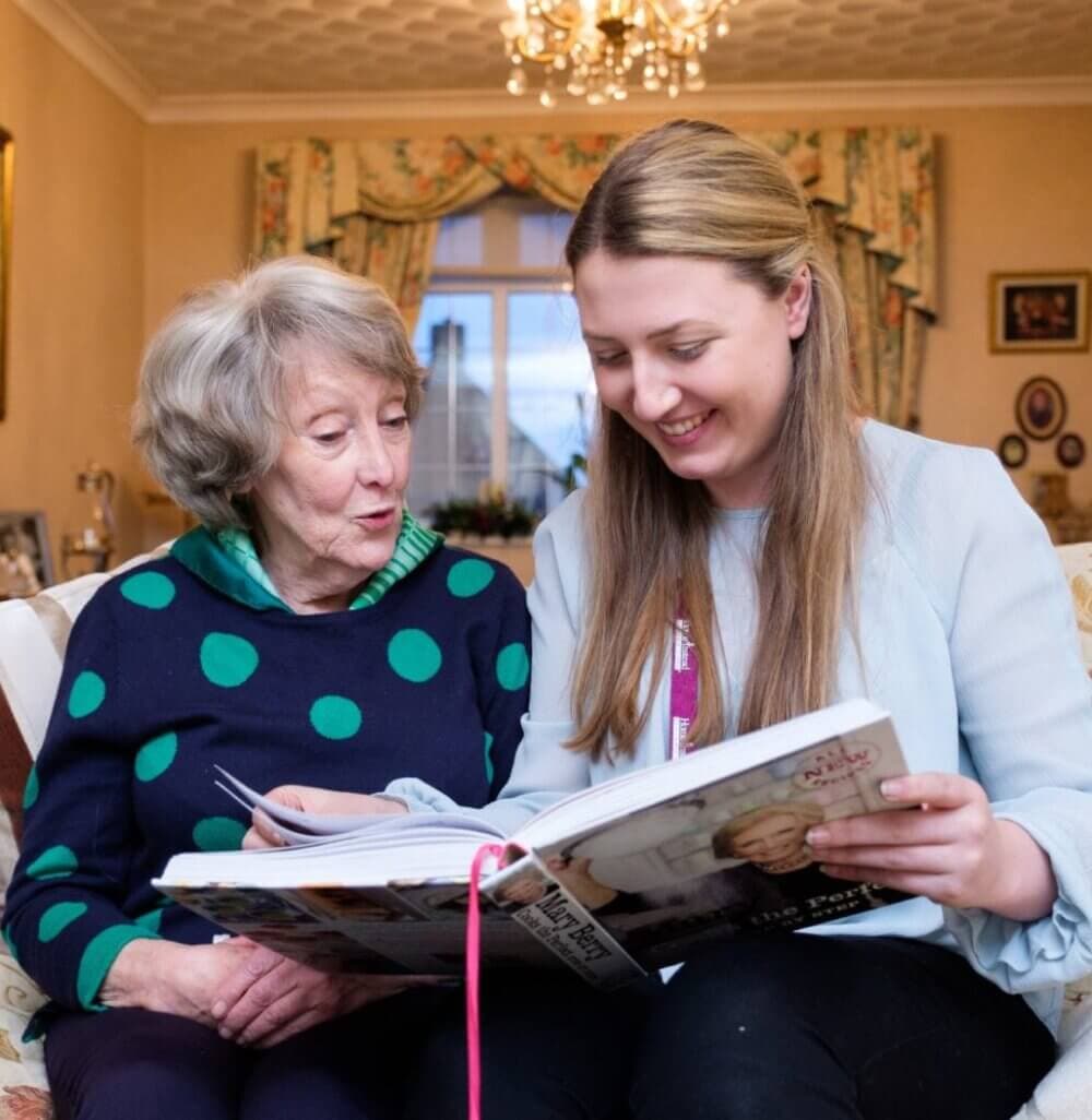 An elderly woman and a young woman sit on a couch, smiling and looking at an open book together in a cozy living room. - Home Instead