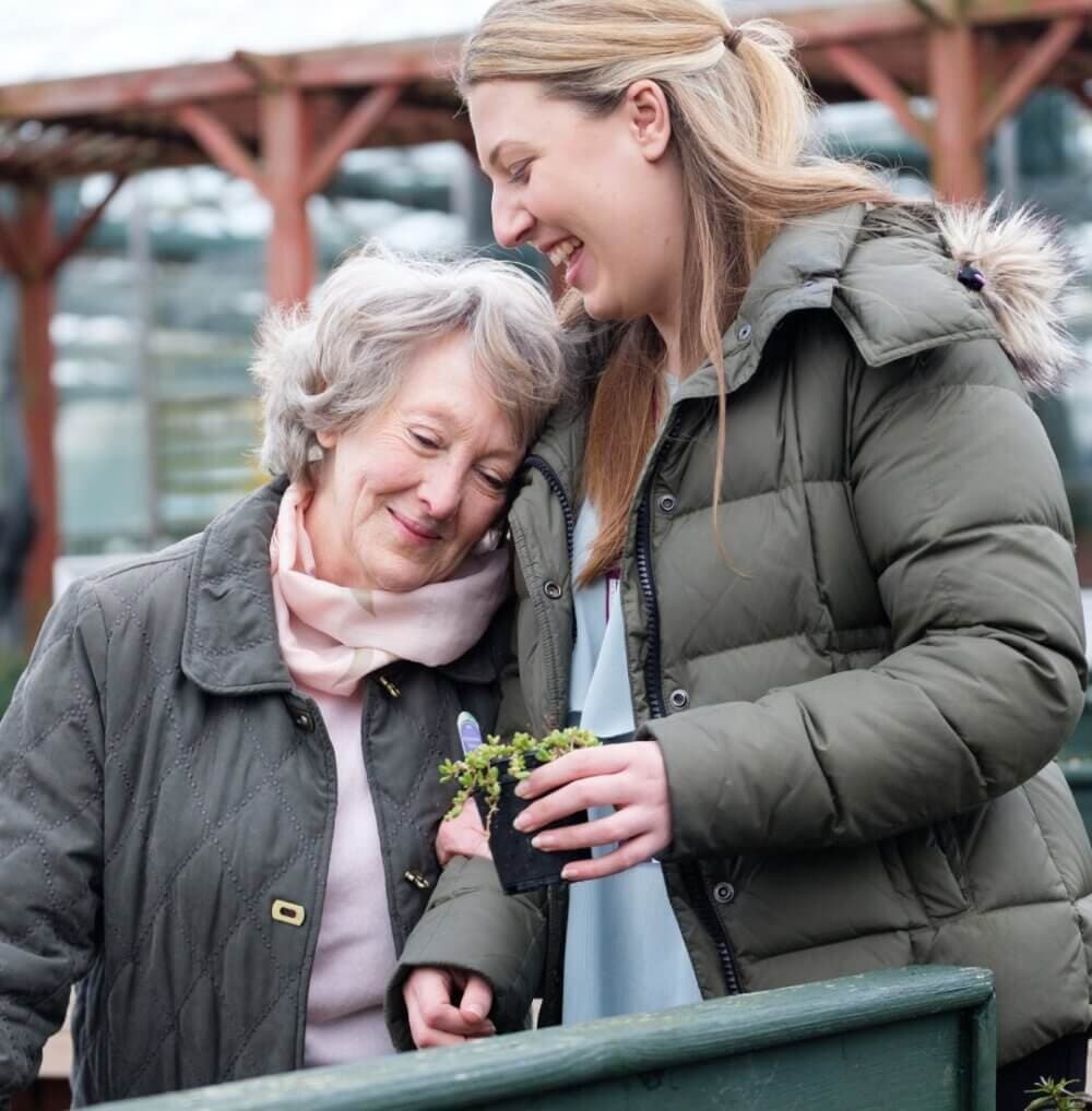 A young woman and an older woman smile and hold a small potted plant together outdoors. - Home Instead
