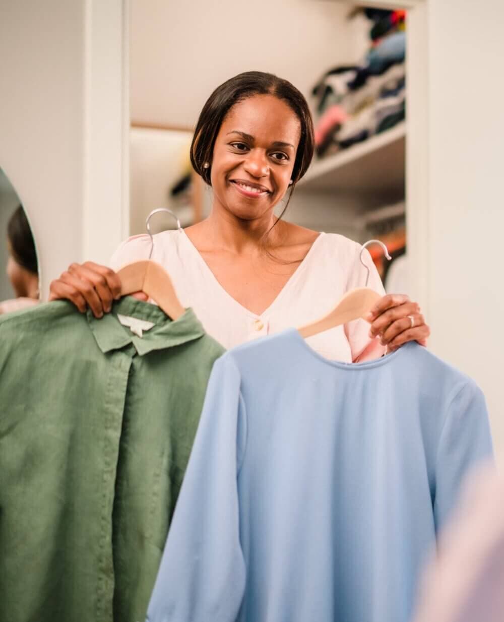 Woman smiling, holding a green shirt on a hanger in one hand and a blue blouse on a second hanger in the other hand. - Home Instead