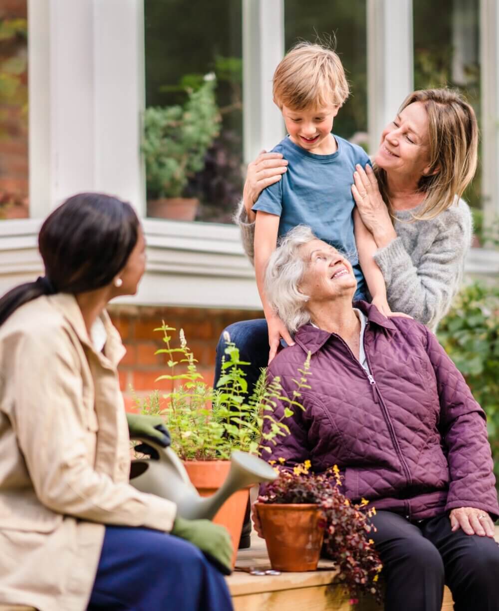 Three generations enjoy a garden: an older woman, two younger women, and a child smiling together on the patio steps. - Home Instead Bournemouth & Christchurch