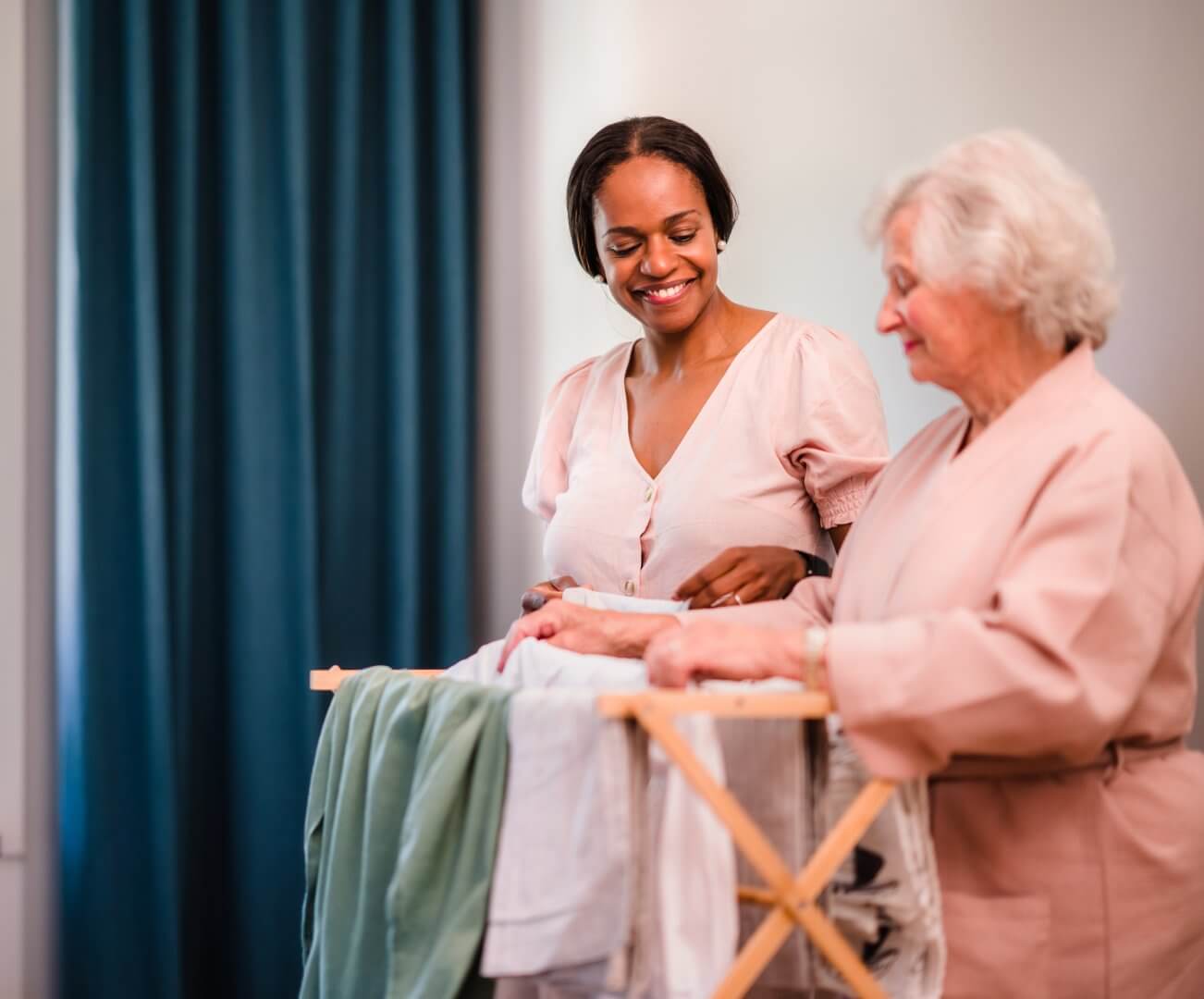 Two women, one younger and one elderly, folding laundry together while smiling in a brightly lit room. - Home Instead