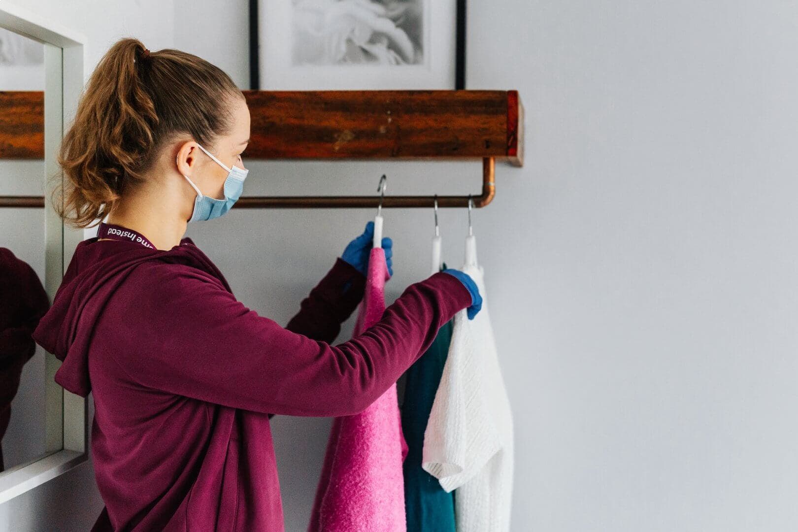 A woman wearing a mask hangs clothes on a rack, handling them with blue gloves. - Home Instead