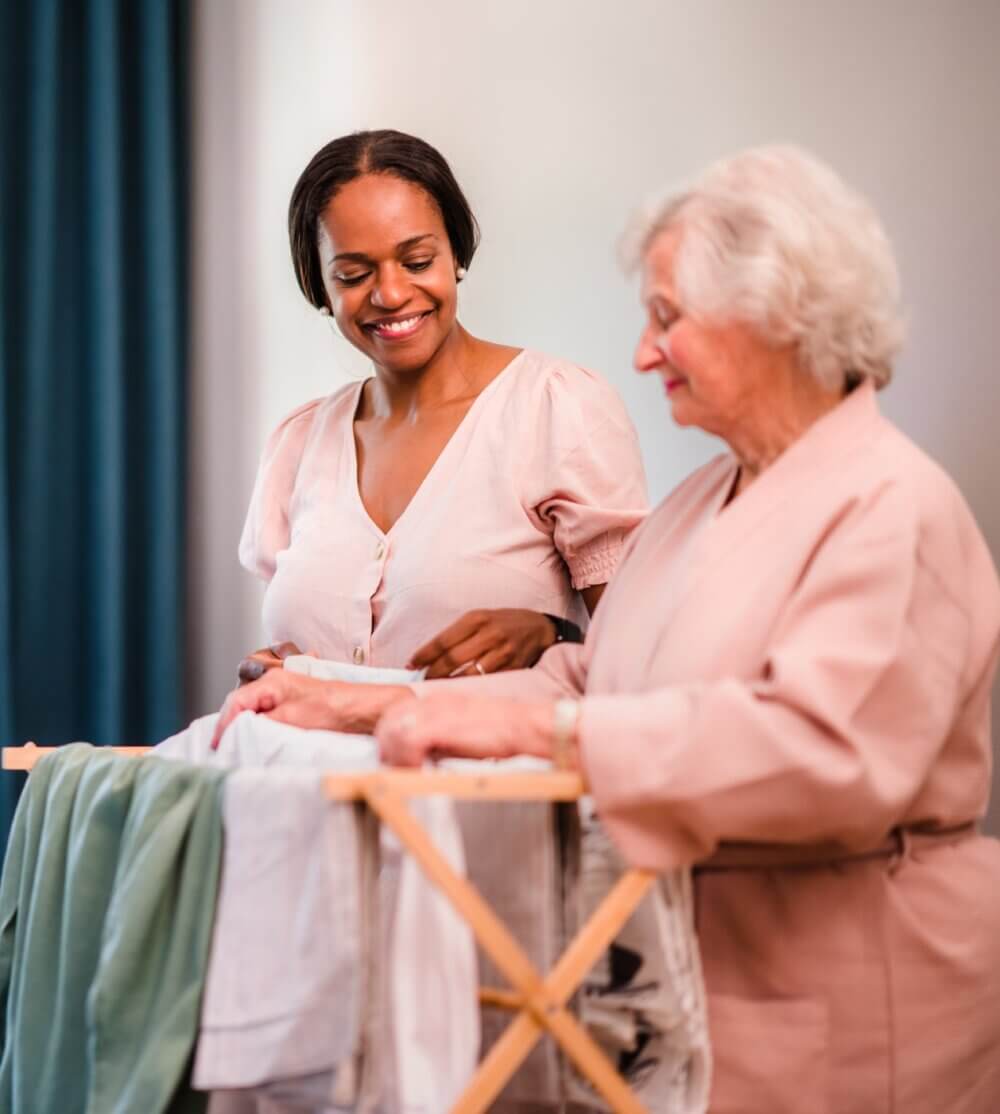 A young woman helps an elderly woman fold laundry together, both smiling and standing by a laundry basket. - Home Instead