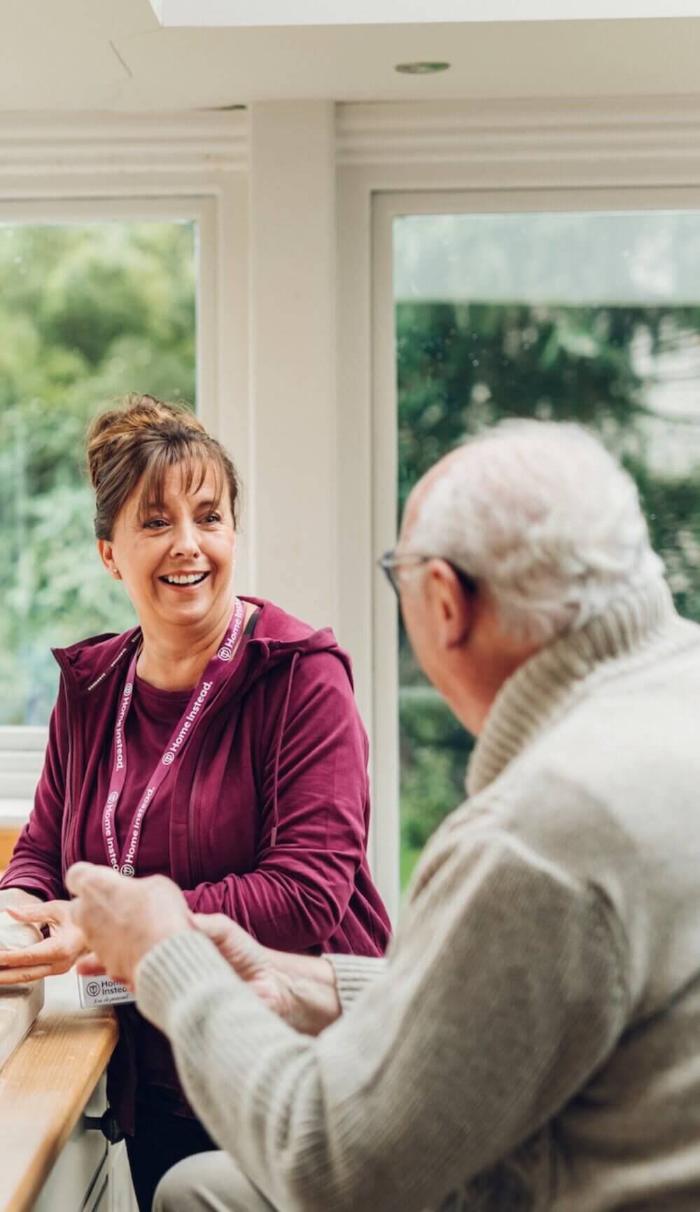 A Care Professional in a burgundy sweater talks and smiles with an elderly man at a table in a bright room with large windows. - Home Instead Bournemouth & Christchurch