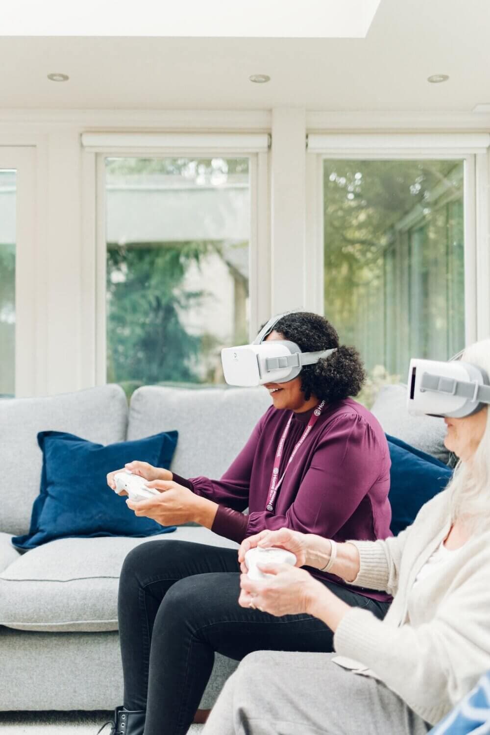Two people using virtual reality headsets and controllers while sitting on a couch in a bright room. - Home Instead