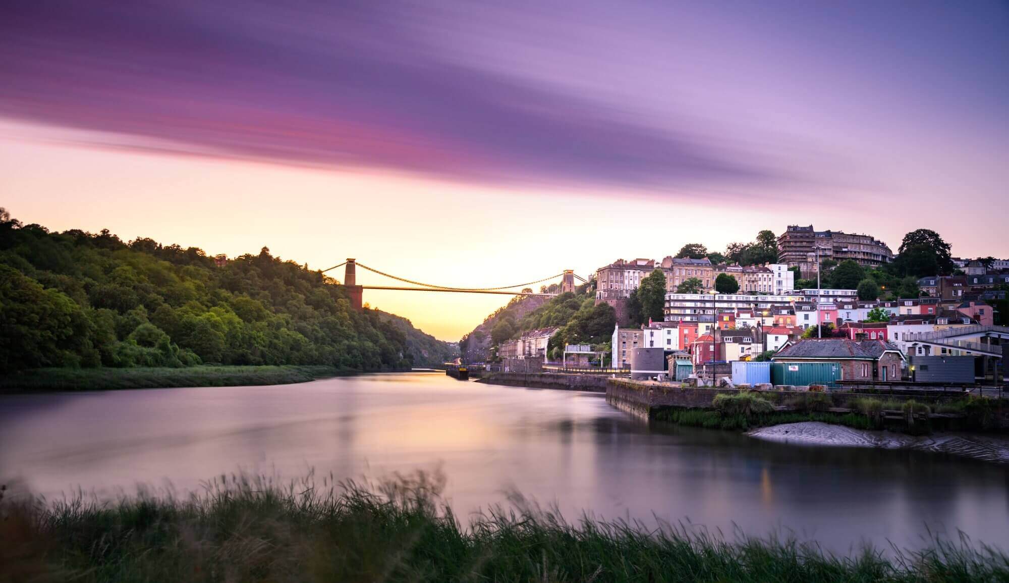A river with a suspension bridge, colorful houses, and lush greenery under a vibrant purple and orange sky at dusk. - Home Instead