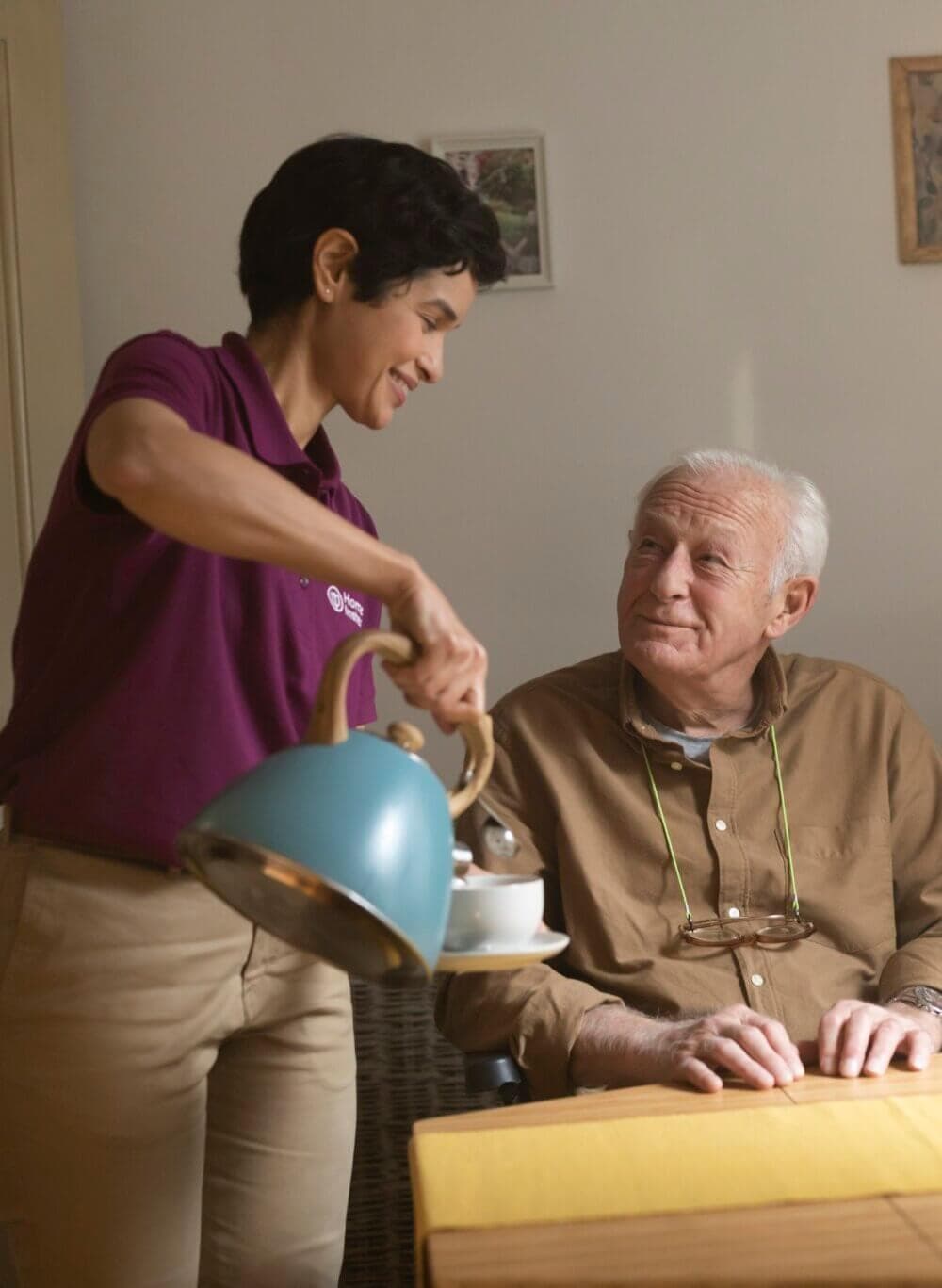A woman pouring tea for an older man sitting at a table, both smiling warmly at each other. - Home Instead