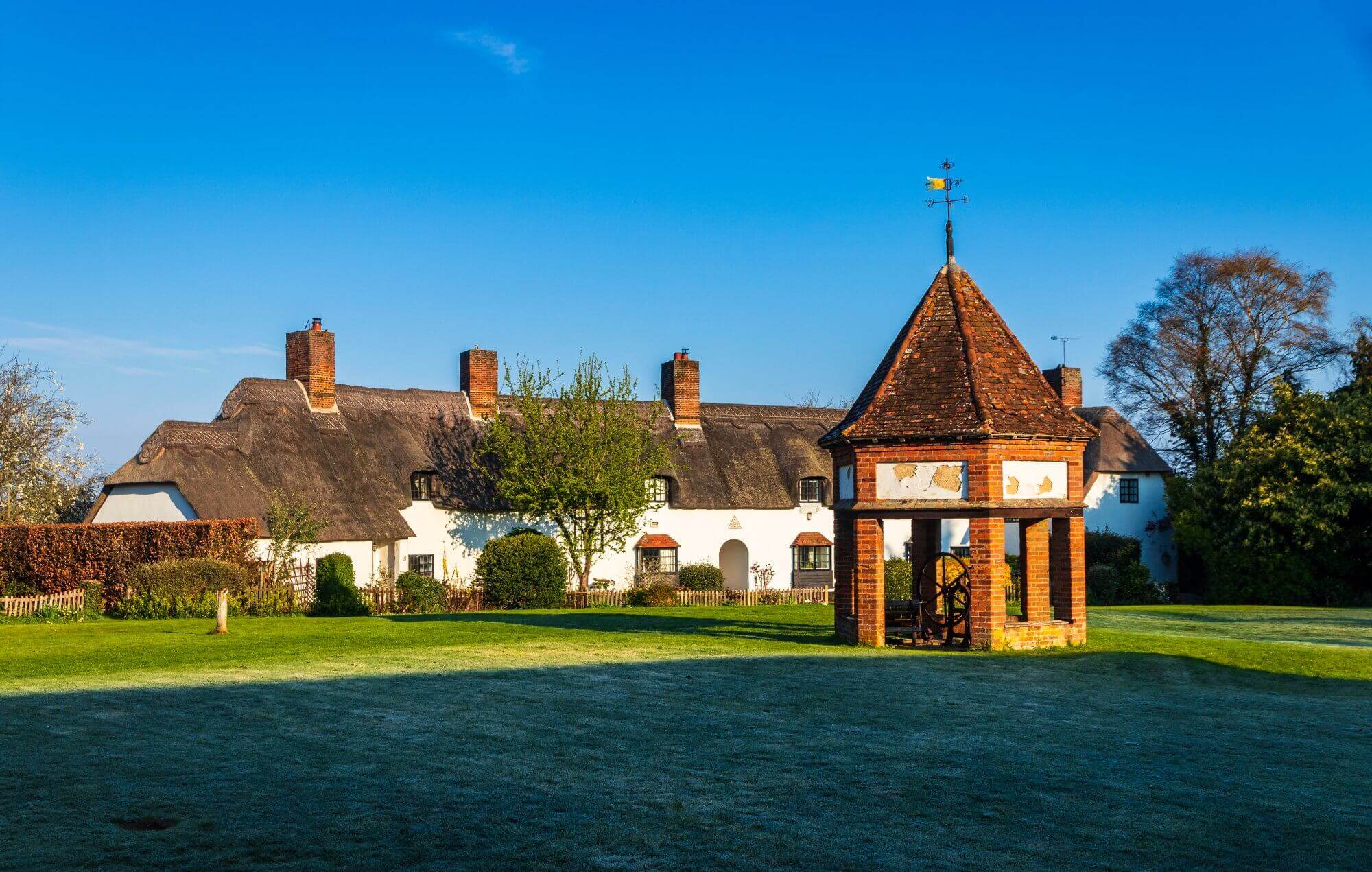 Scenic view of thatched cottages with a brick gazebo and green lawn under a clear blue sky. - Home Instead