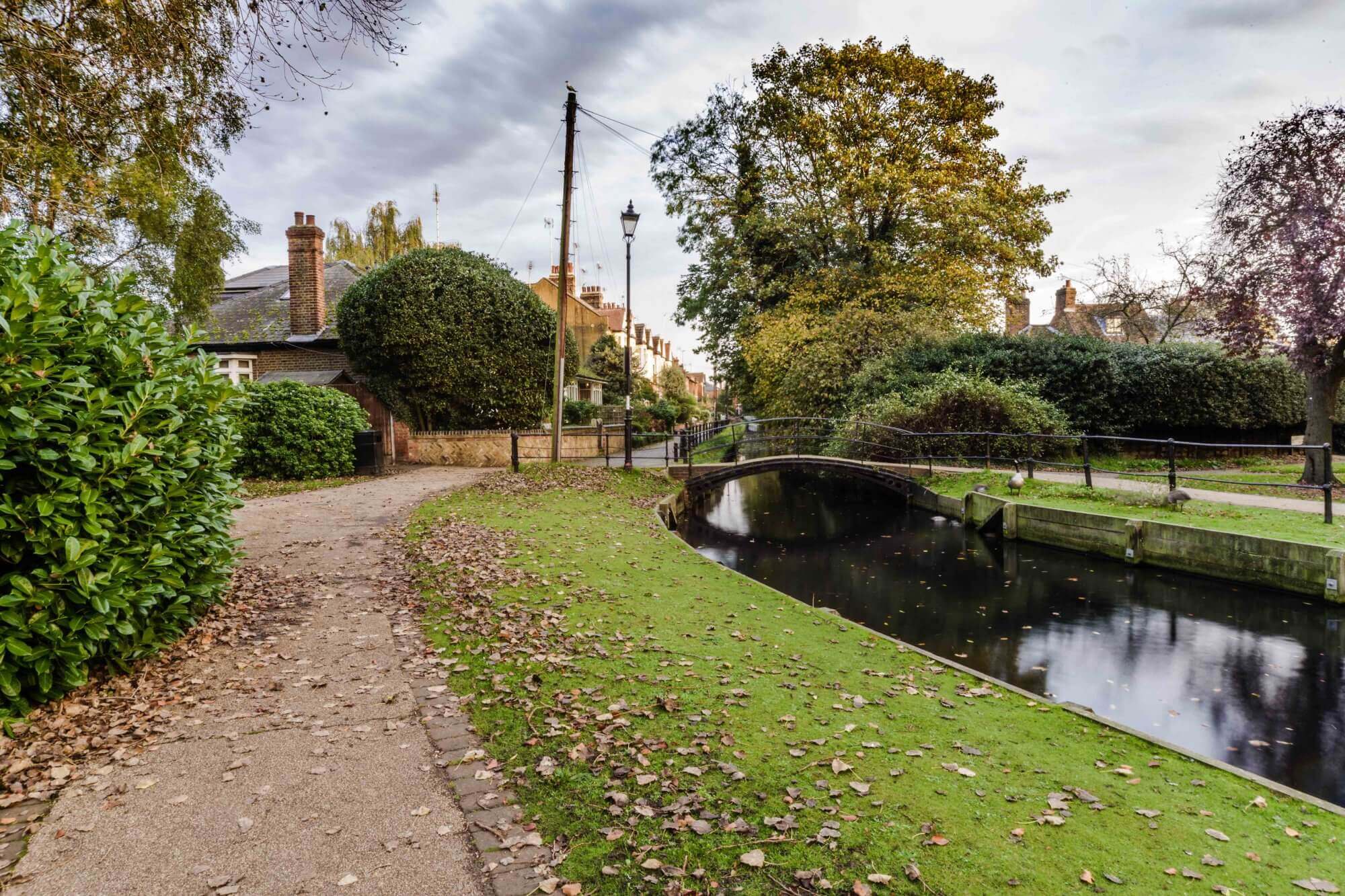A serene park path running alongside a calm stream with a small arched bridge and adjacent houses on a cloudy day. - Home Instead