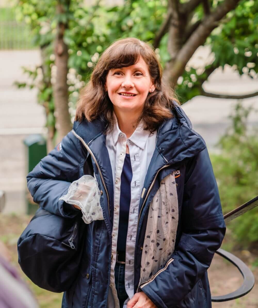 A woman in a blue jacket smiling outdoors, holding a black bag and a transparent plastic item. - Home Instead