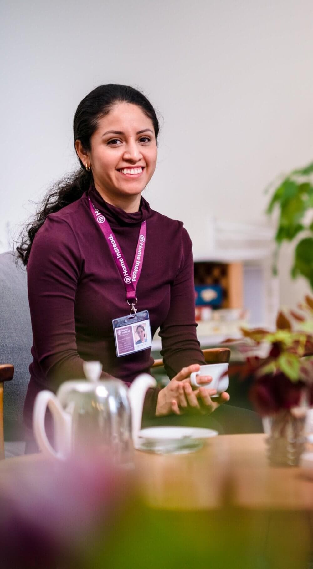 A woman in a maroon top and lanyard sits in a cozy setting holding a cup, with a teapot and plants nearby. - Home Instead