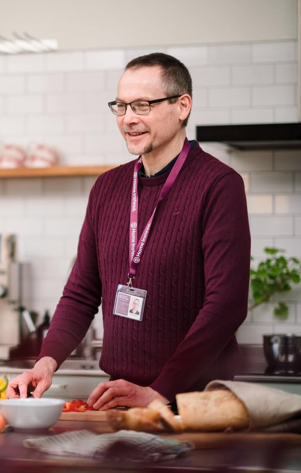 A person in a burgundy sweater and glasses smiles while preparing food in a kitchen. - Home Instead