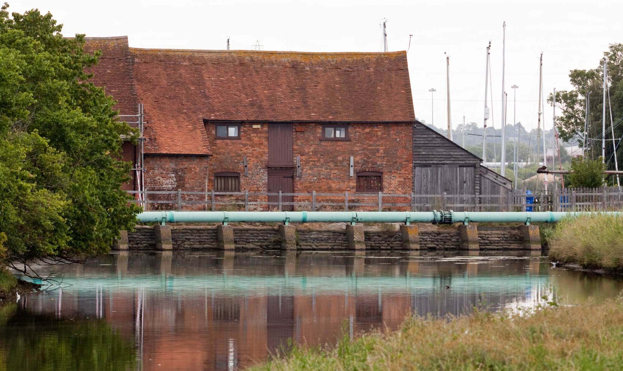 Brick building with a tiled roof by a river, featuring a large pipe and surrounded by trees and boats in the background. - Home Instead