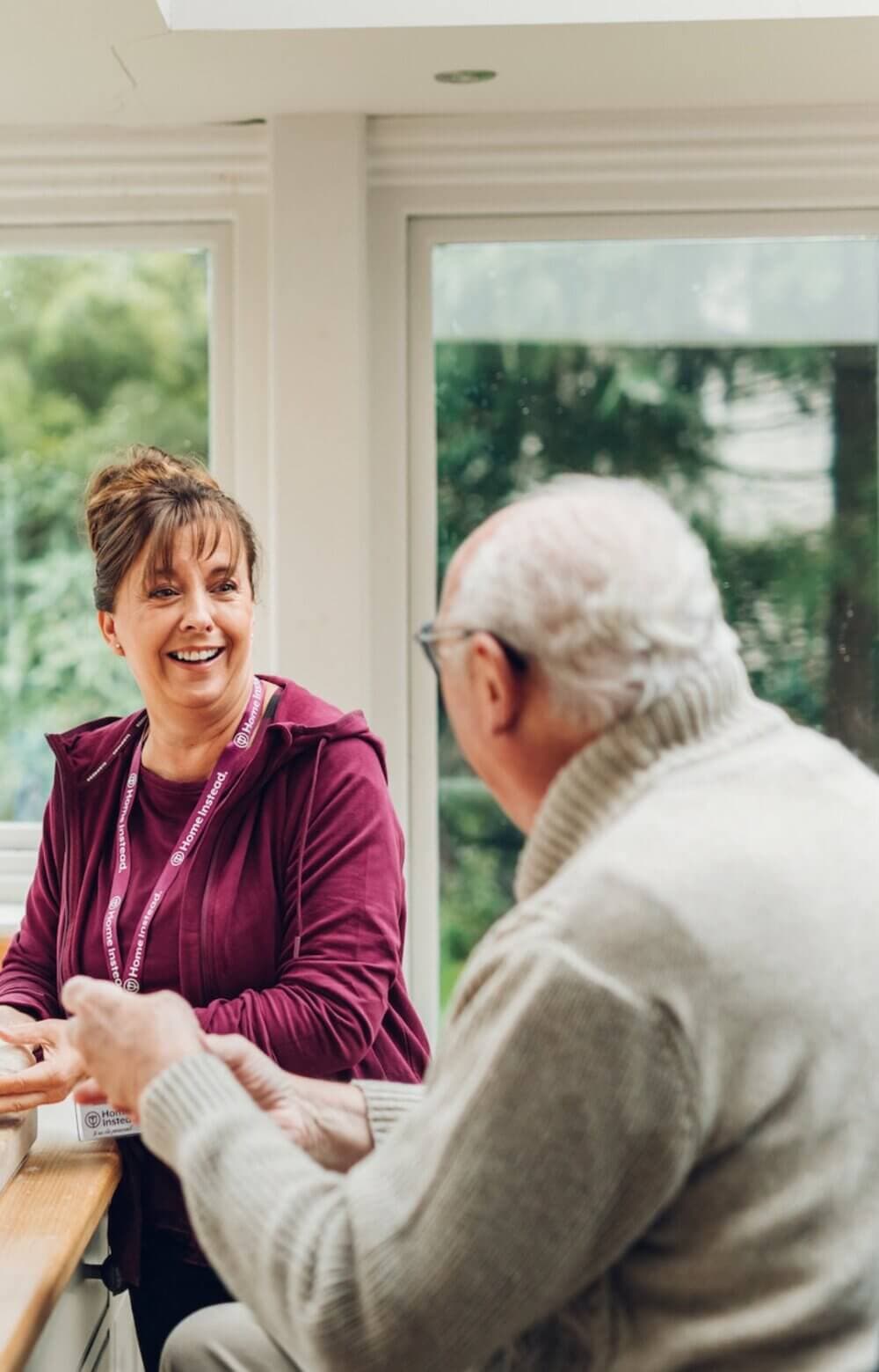 A woman in a maroon hoodie smiles while talking to an older man in a white sweater at a table. - Home Instead Poole