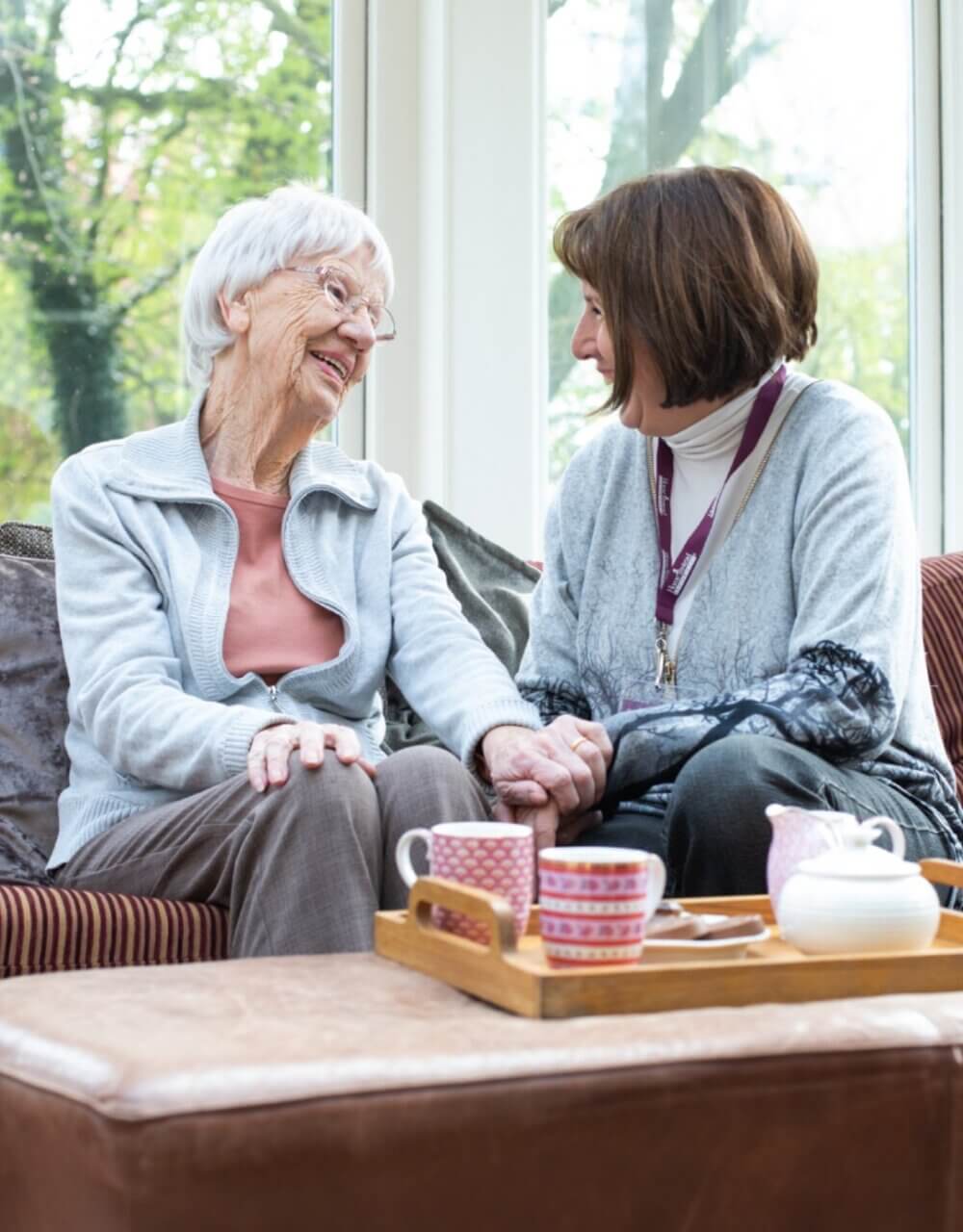An elderly woman and a younger woman sit on a couch, smiling and holding hands, with tea cups on a tray in front of them. - Home Instead