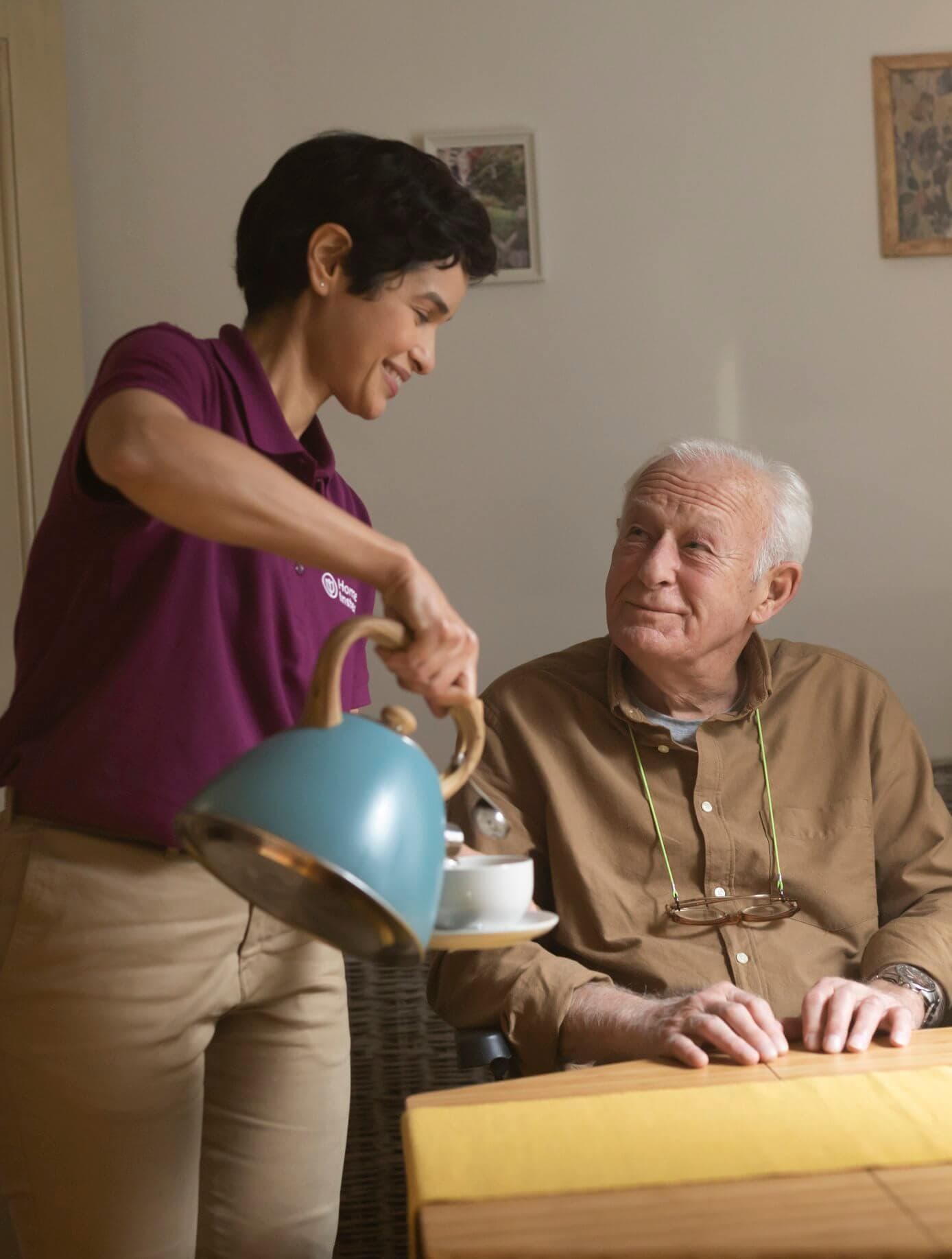 Caregiver in a purple shirt pours tea for an elderly man sitting at a table, both smiling warmly. - Home Instead