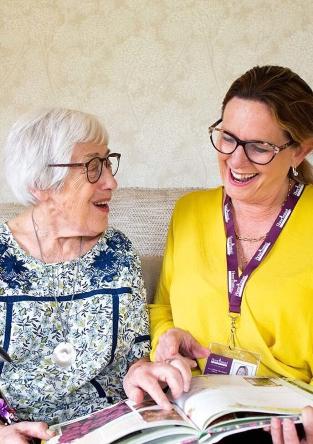 An elderly woman and a caregiver with glasses smile while looking at a book together. - Home Instead