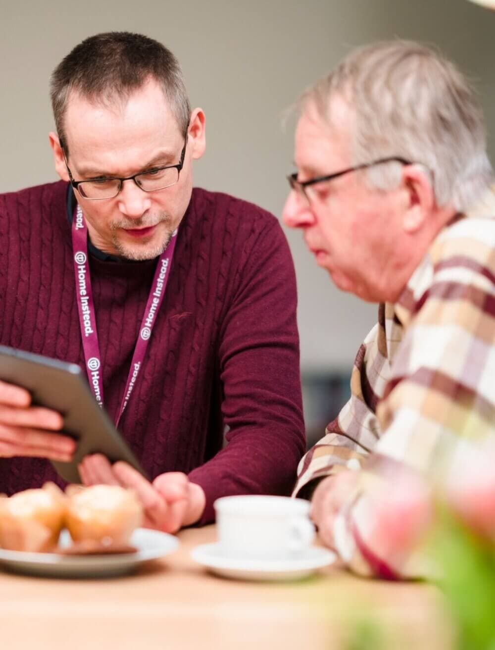 A man with glasses shows a tablet to an older man over a table with pastries and a coffee cup. - Home Instead