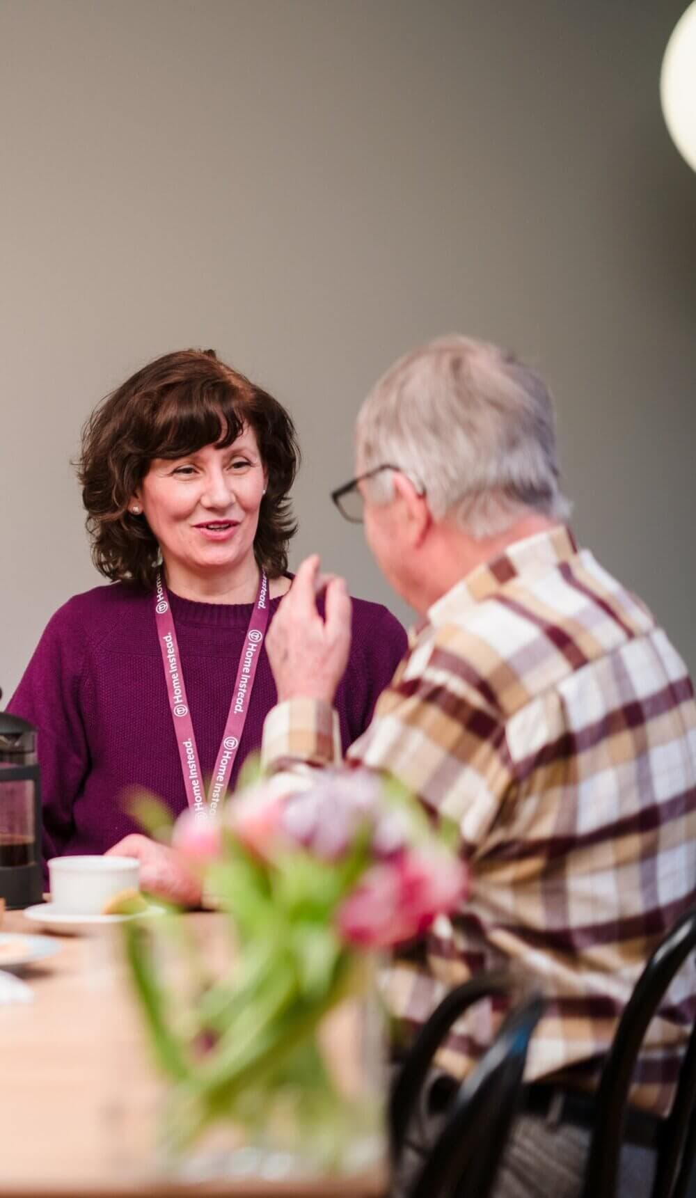 A woman and an older man are talking at a table with flowers. She is wearing a lanyard; he is in a plaid shirt. - Home Instead