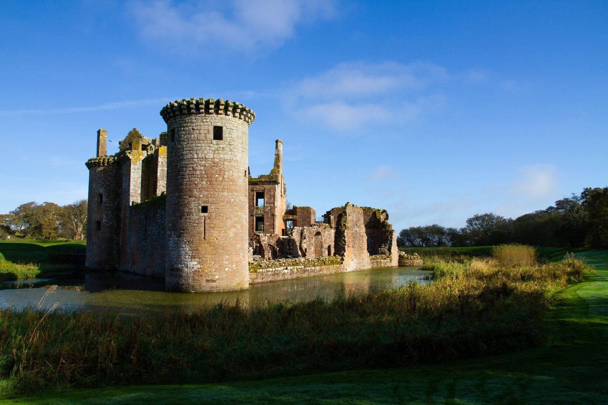 Medieval stone castle with partially ruined towers and walls, surrounded by a moat and lush greenery under a bright blue sky. - Home Instead