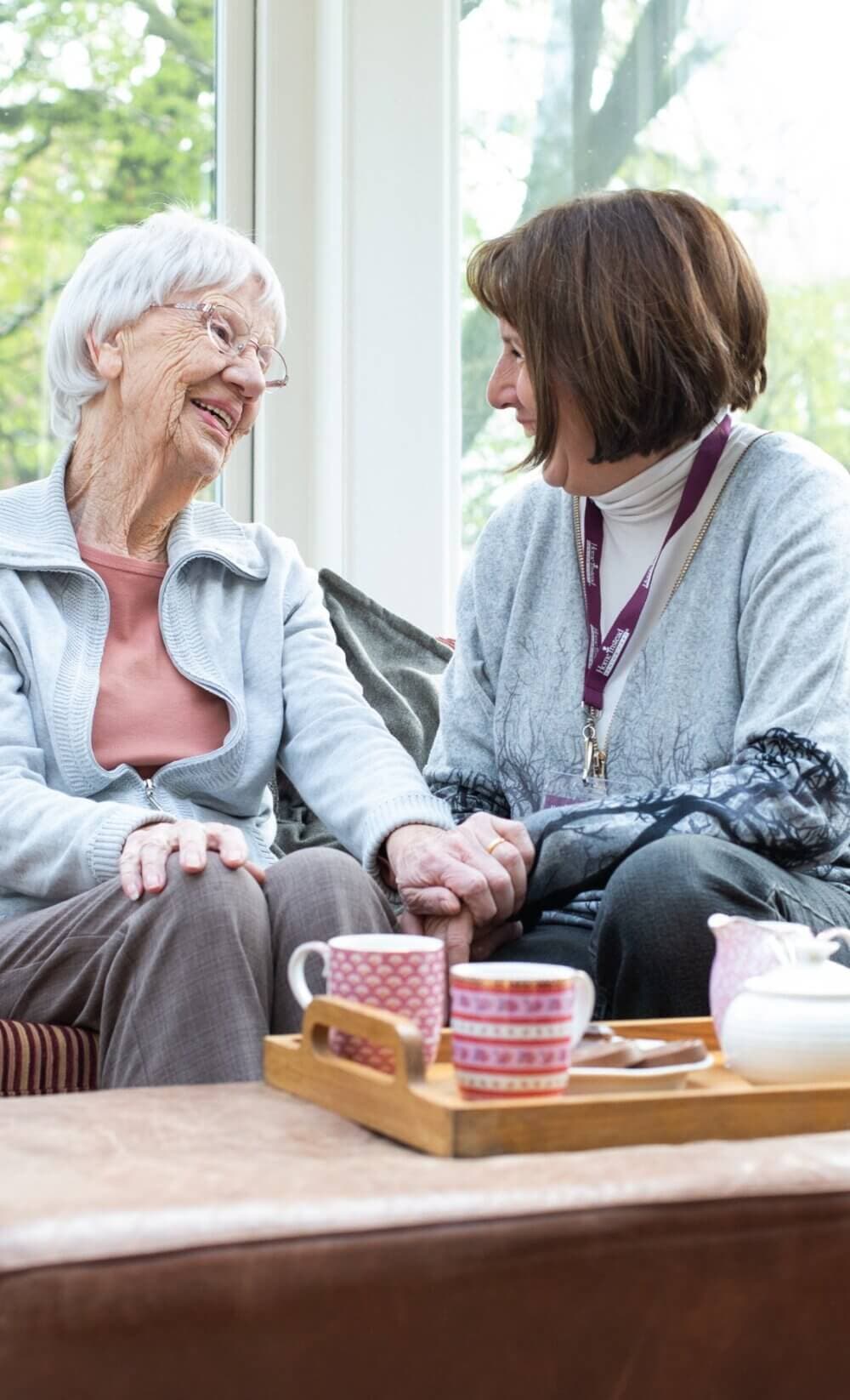 An elderly woman and a younger woman share a smile and hold hands, seated by a window with tea cups in front of them. - Home Instead Southampton
