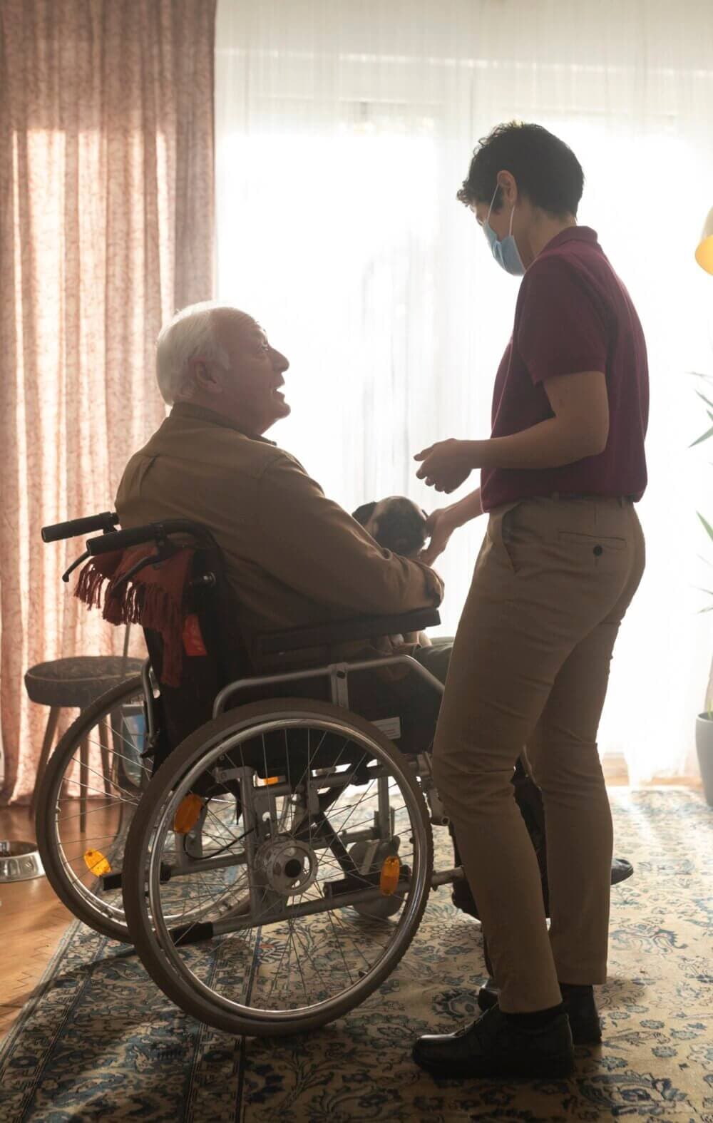 An elderly man in a wheelchair and a caregiver wearing a mask interact warmly in a sunlit room. - Home Instead