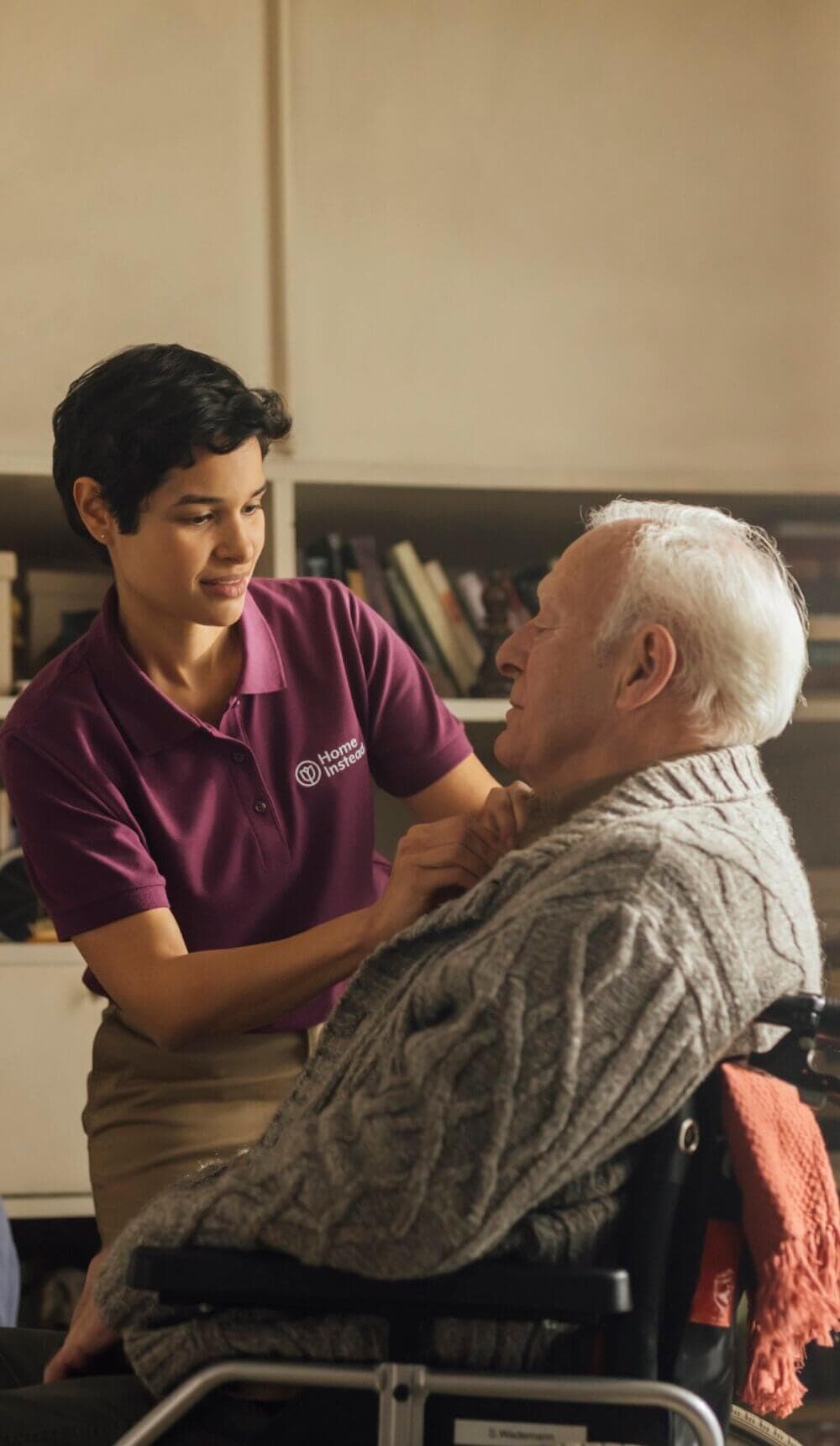Caregiver assists an elderly man in a wheelchair, adjusting his sweater in a cozy room with books on shelves in the background. - Home Instead