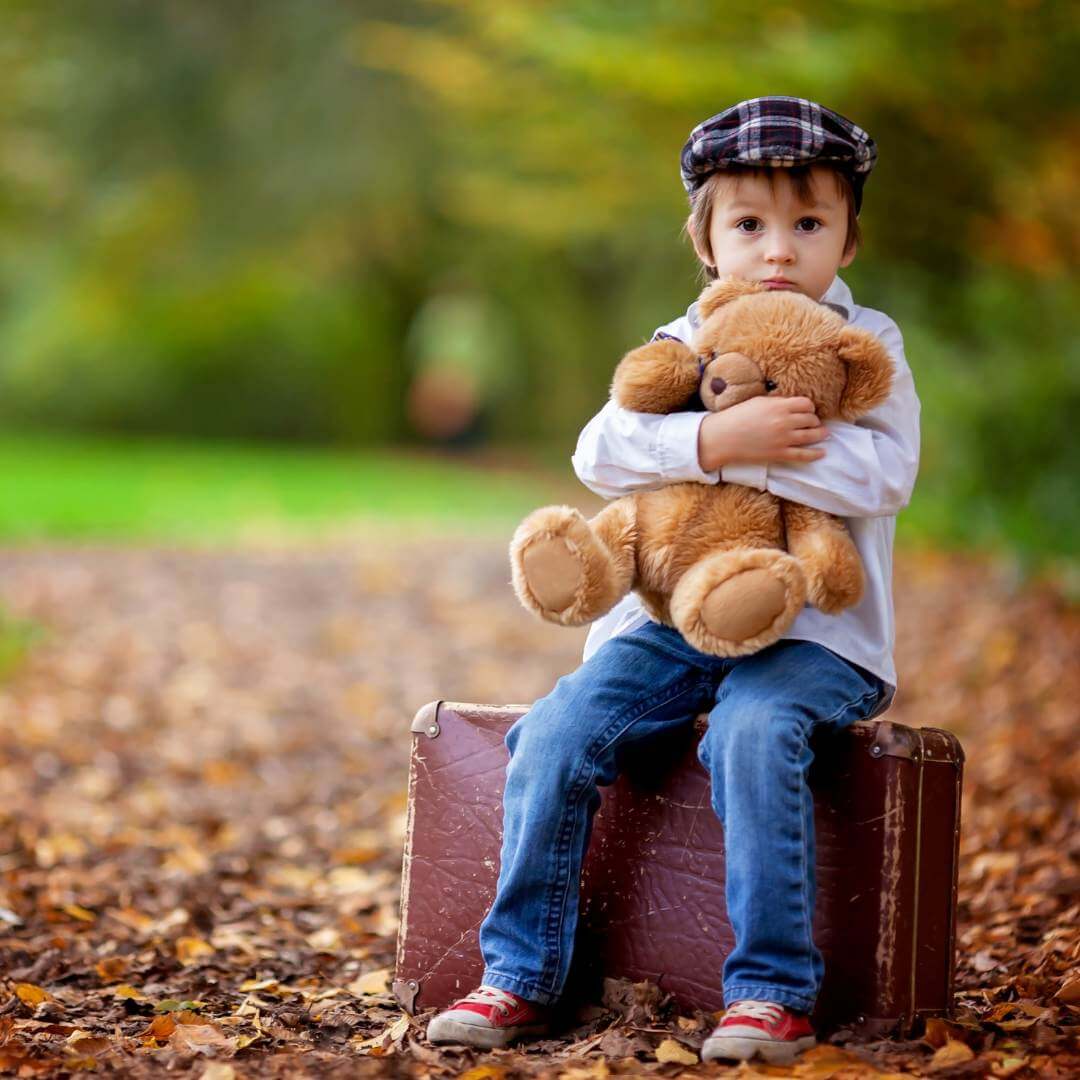 Young boy sitting on a suitcase, hugging a teddy bear amidst an autumn landscape with leaves on the ground. - Home Instead