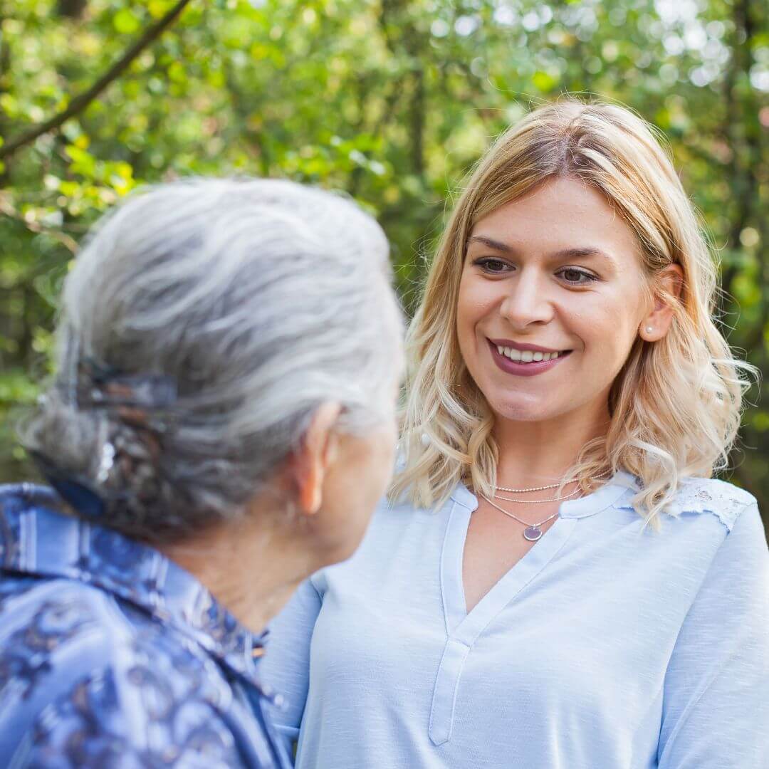 Two women, one elderly and one young, smiling at each other in an outdoor setting with greenery in the background. - Home Instead