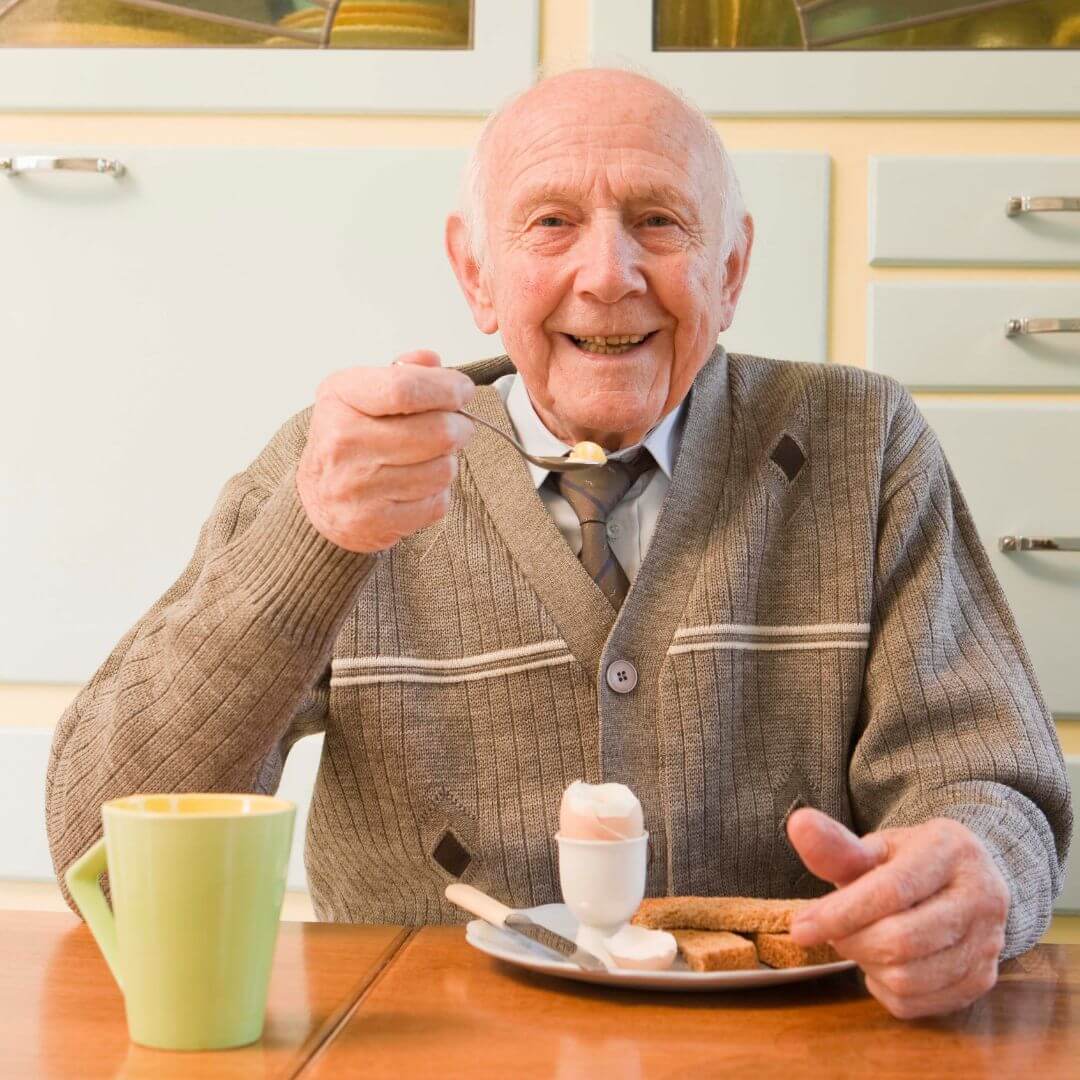 An elderly man smiles while eating an egg at the table, with a green mug and toast in the foreground. - Home Instead
