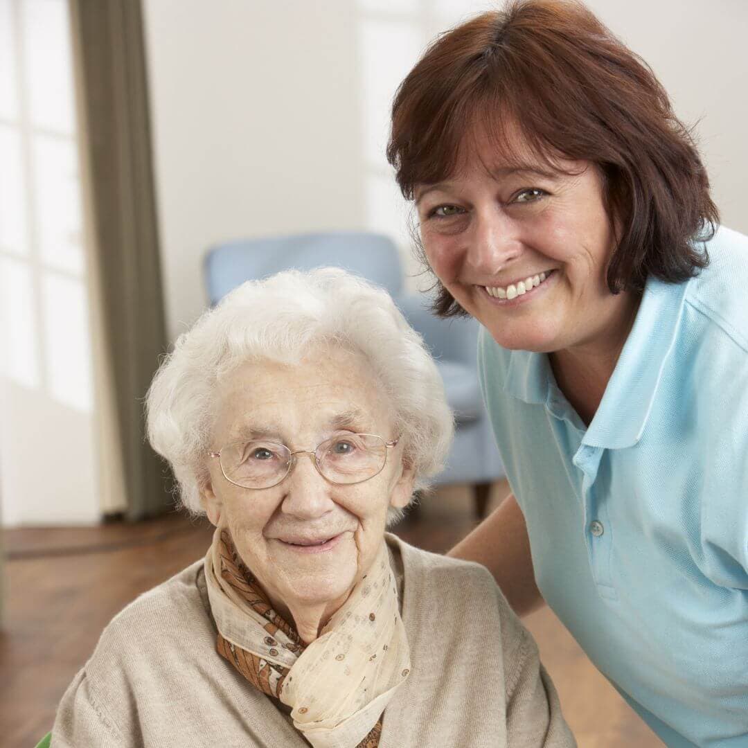 An elderly woman with glasses and a scarf smiles while sitting next to a woman with short brown hair in a blue shirt. - Home Instead