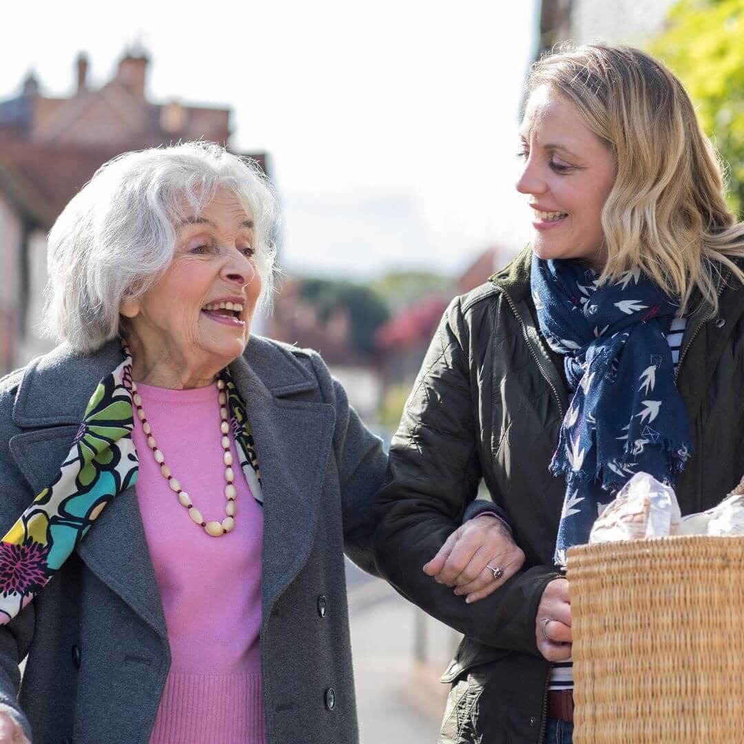 Elderly woman in colorful scarf smiles while walking arm-in-arm with younger woman carrying a basket. - Home Instead