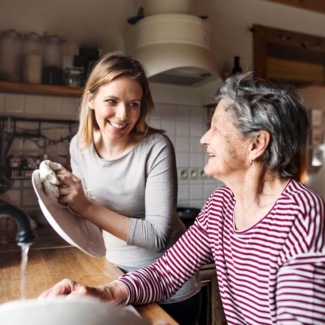 A woman washes dishes while smiling and talking to an elderly woman in a cozy kitchen. - Home Instead