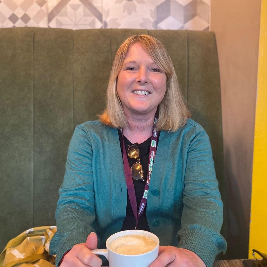 A woman with blonde hair is smiling, sitting in a cafe booth, holding a cup of coffee. She is wearing a teal sweater. - Home Instead