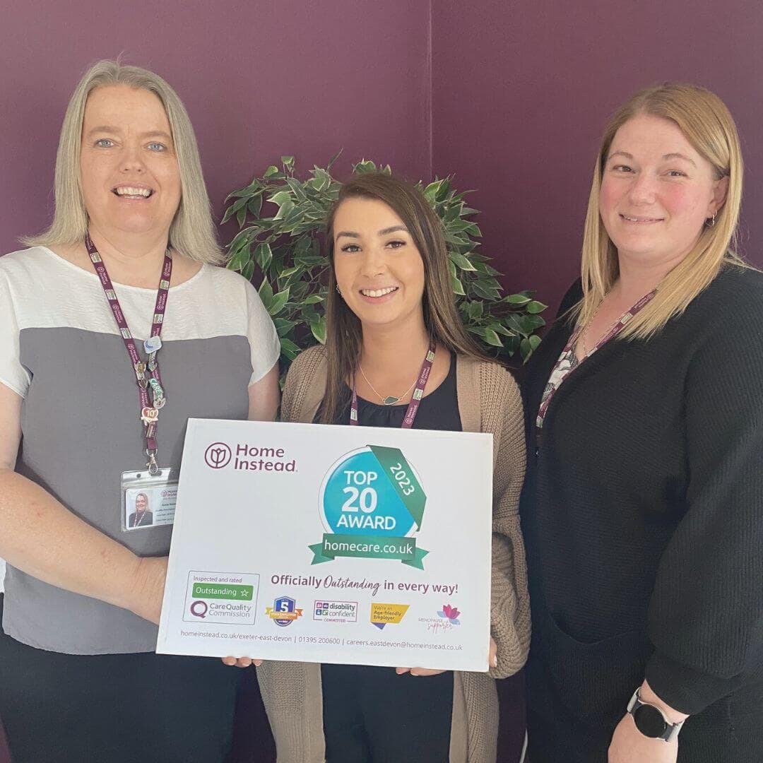 Three women smile while holding a "Top 20 Award" sign from homecare.co.uk for Home Instead, in front of a purple wall. - Home Instead