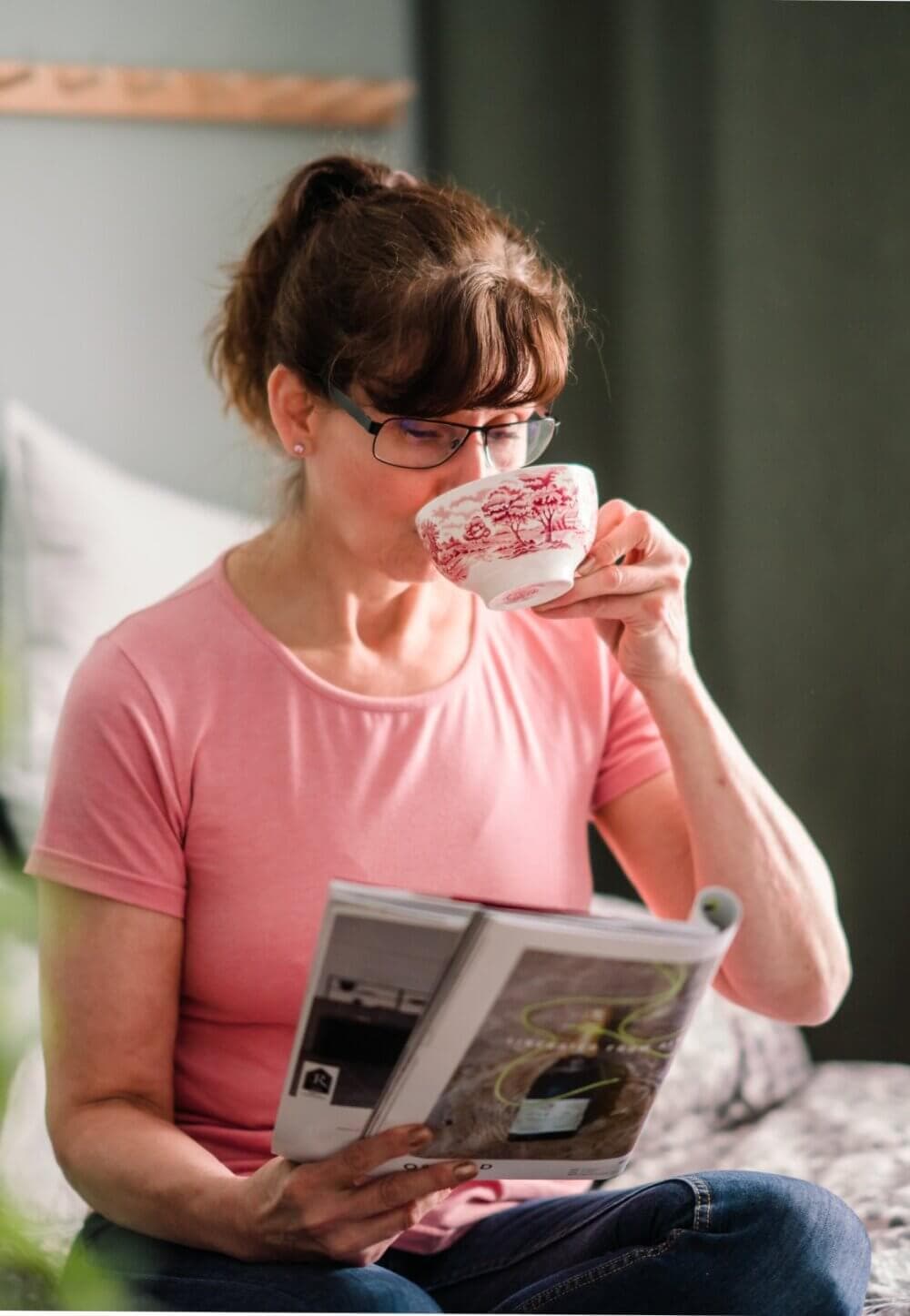 Woman in a pink shirt, wearing glasses, sits on a bed sipping tea and reading a magazine. - Home Instead
