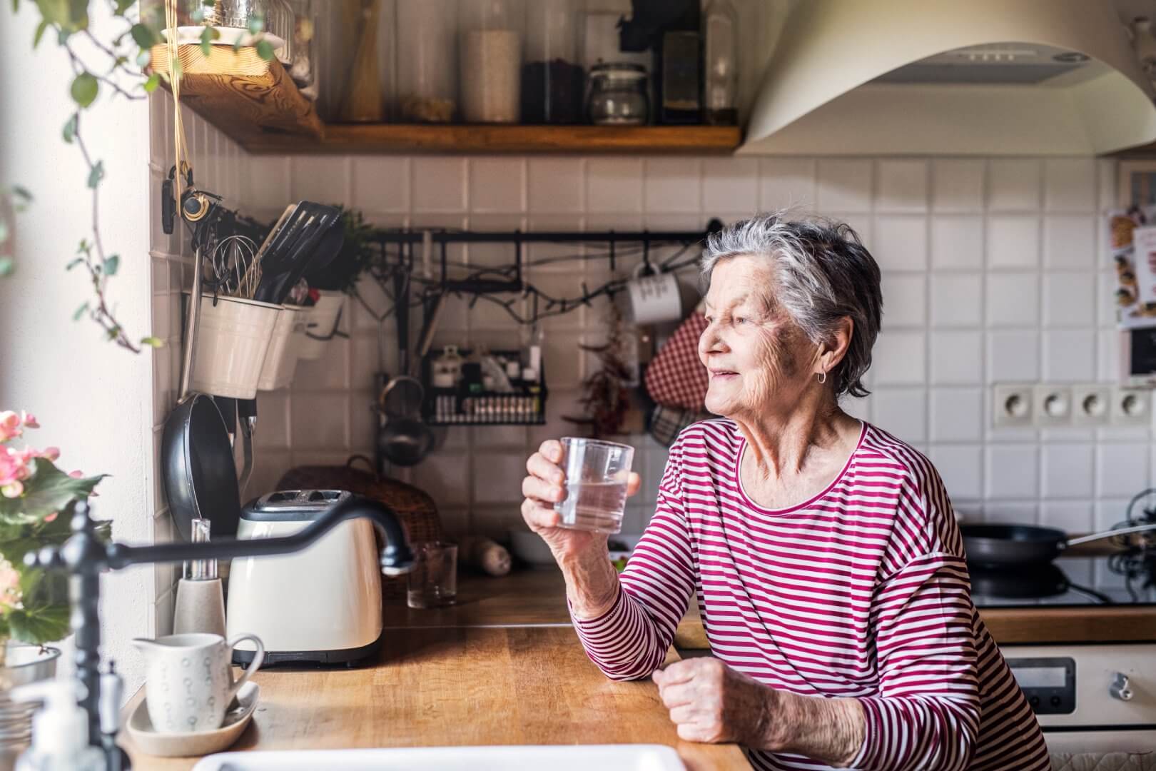 Elderly woman in a striped shirt, holding a glass of water, looks out the window from a cozy kitchen. - Home Instead