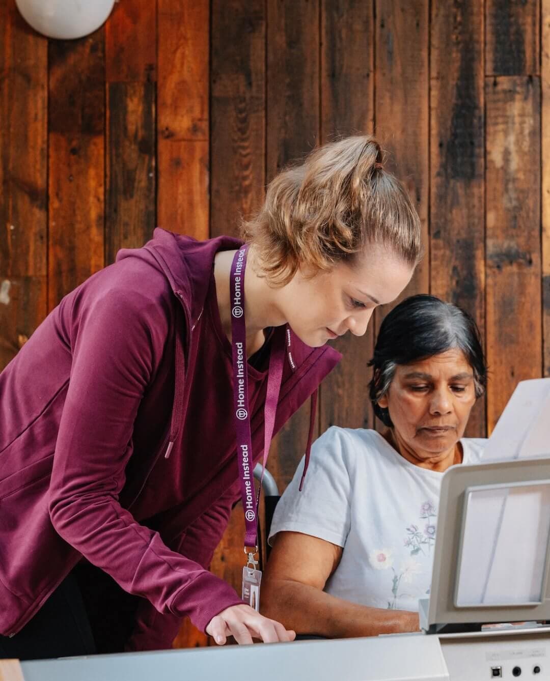 A young woman helps an older woman use a printer in a room with wooden walls. - Home Instead