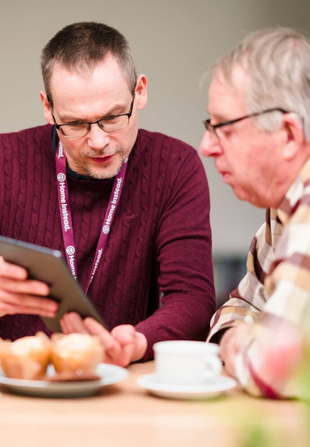 Two men sitting at a table, one showing something on a tablet to the other, with muffins and a coffee cup in view. - Home Instead