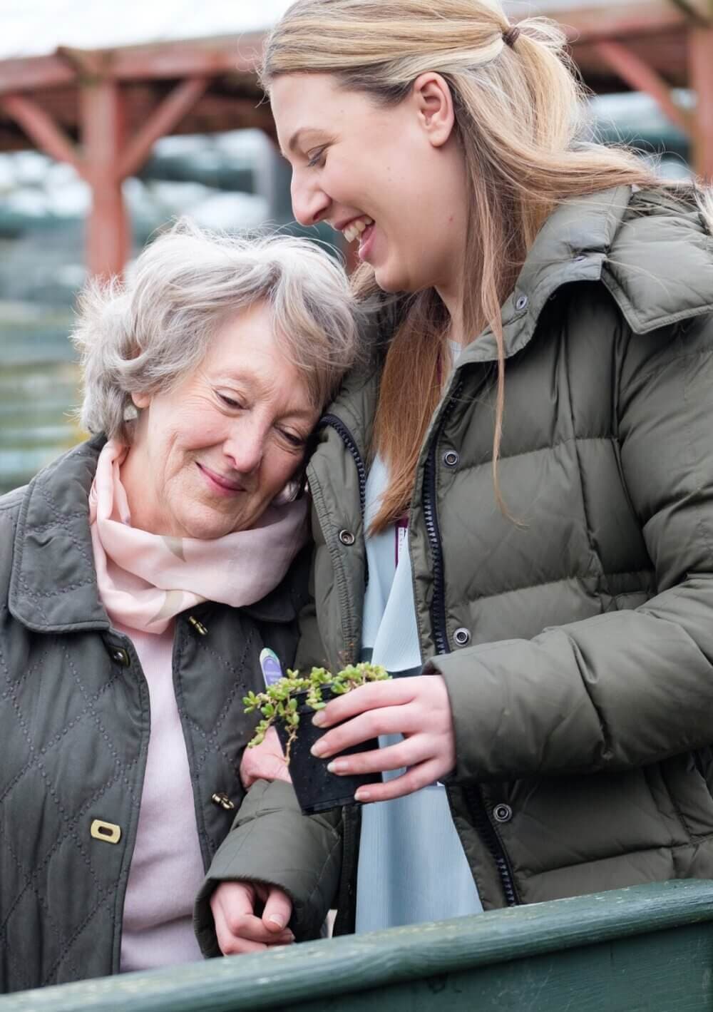 An elderly woman and a younger woman smile and embrace while holding a small potted plant outdoors. - Home Instead