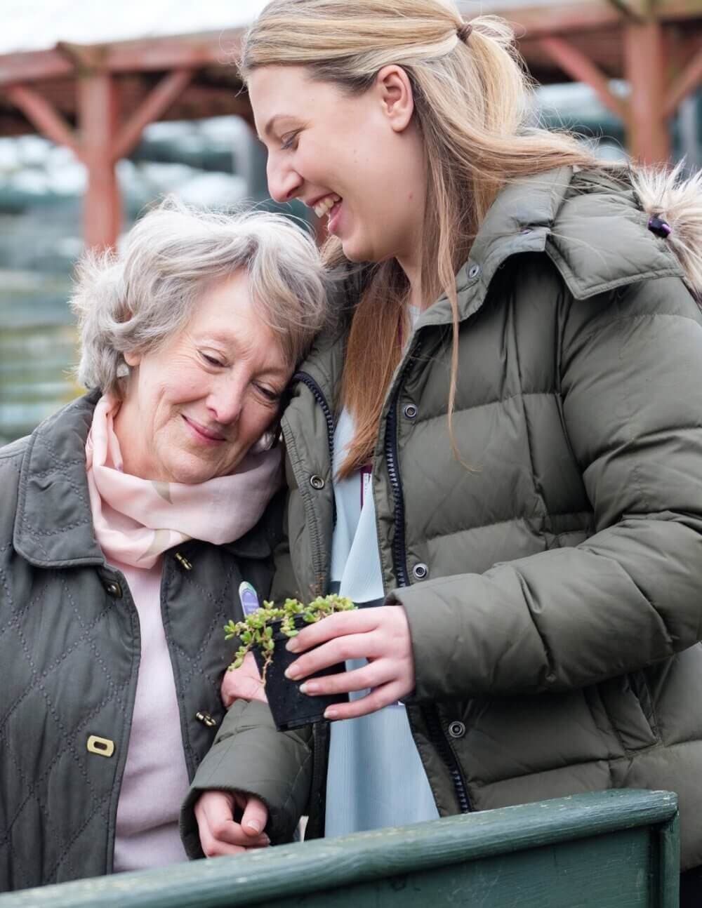 Elderly woman and younger woman smiling together outdoors, holding a small potted plant. - Home Instead