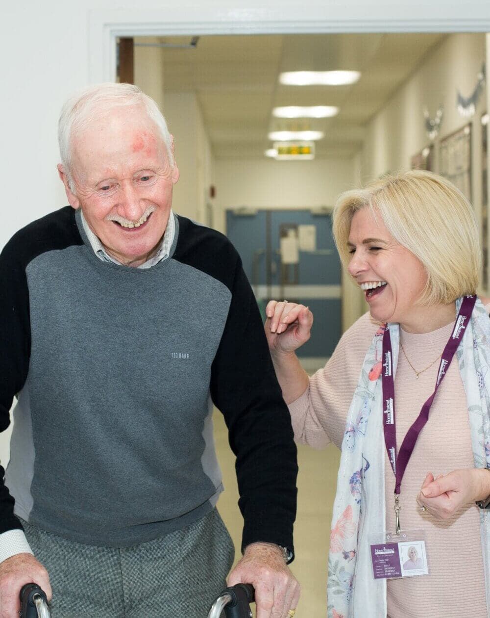 An elderly man with a walker and a woman with a lanyard around her neck laugh together in a hallway. - Home Instead