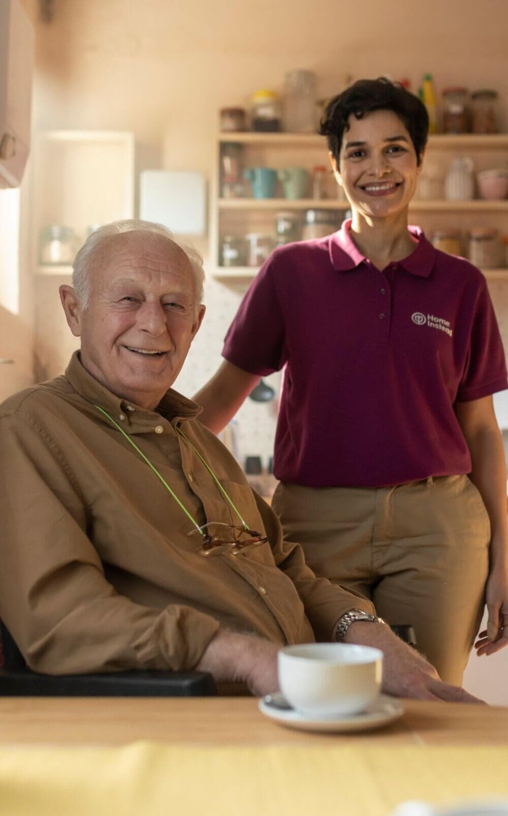 An elderly man sits at a table with a cup, while a smiling caregiver stands beside him in a kitchen. - Home Instead